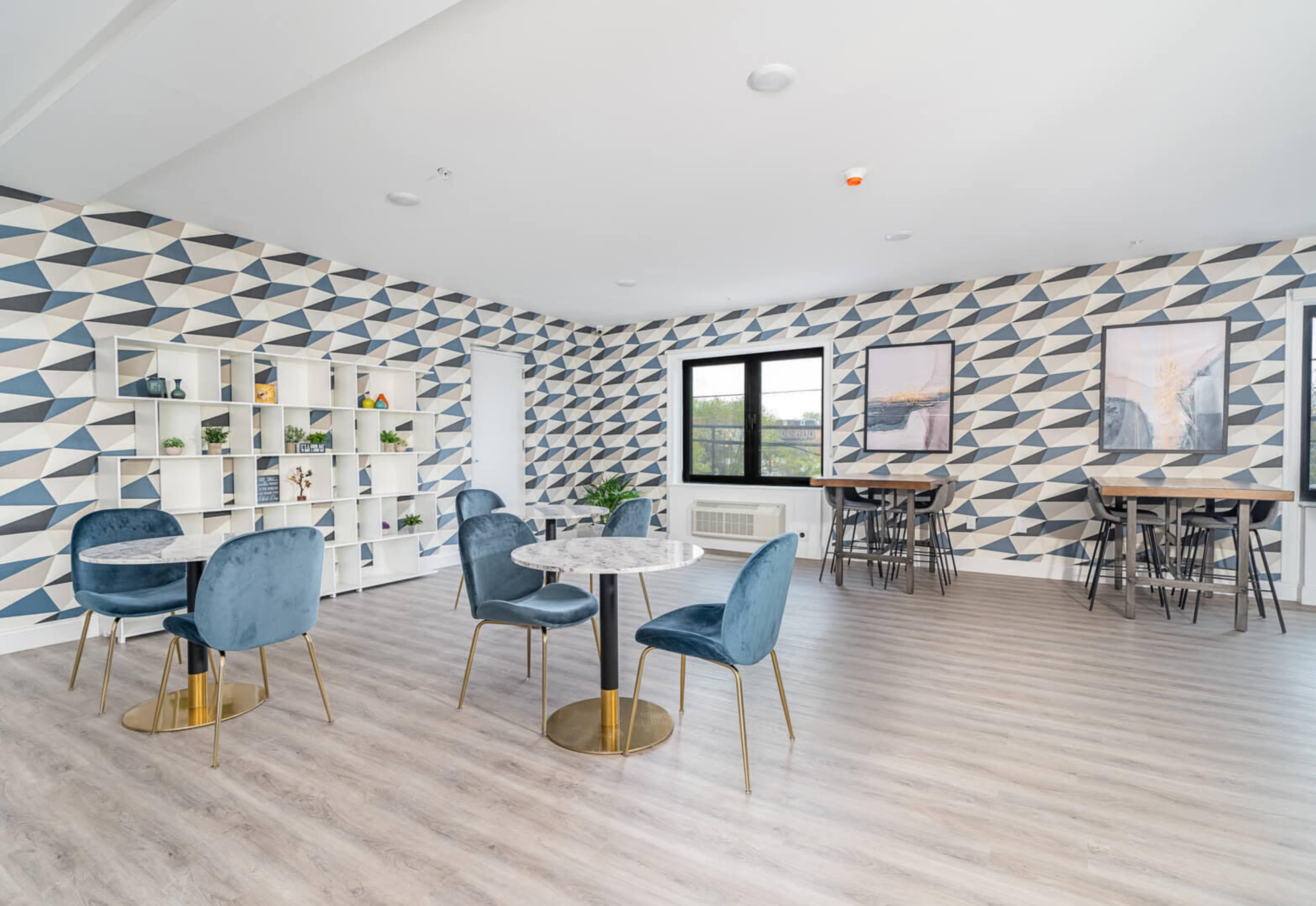 A modern communal room with geometric wallpaper, featuring stylish tables and chairs, and large windows allowing natural light.