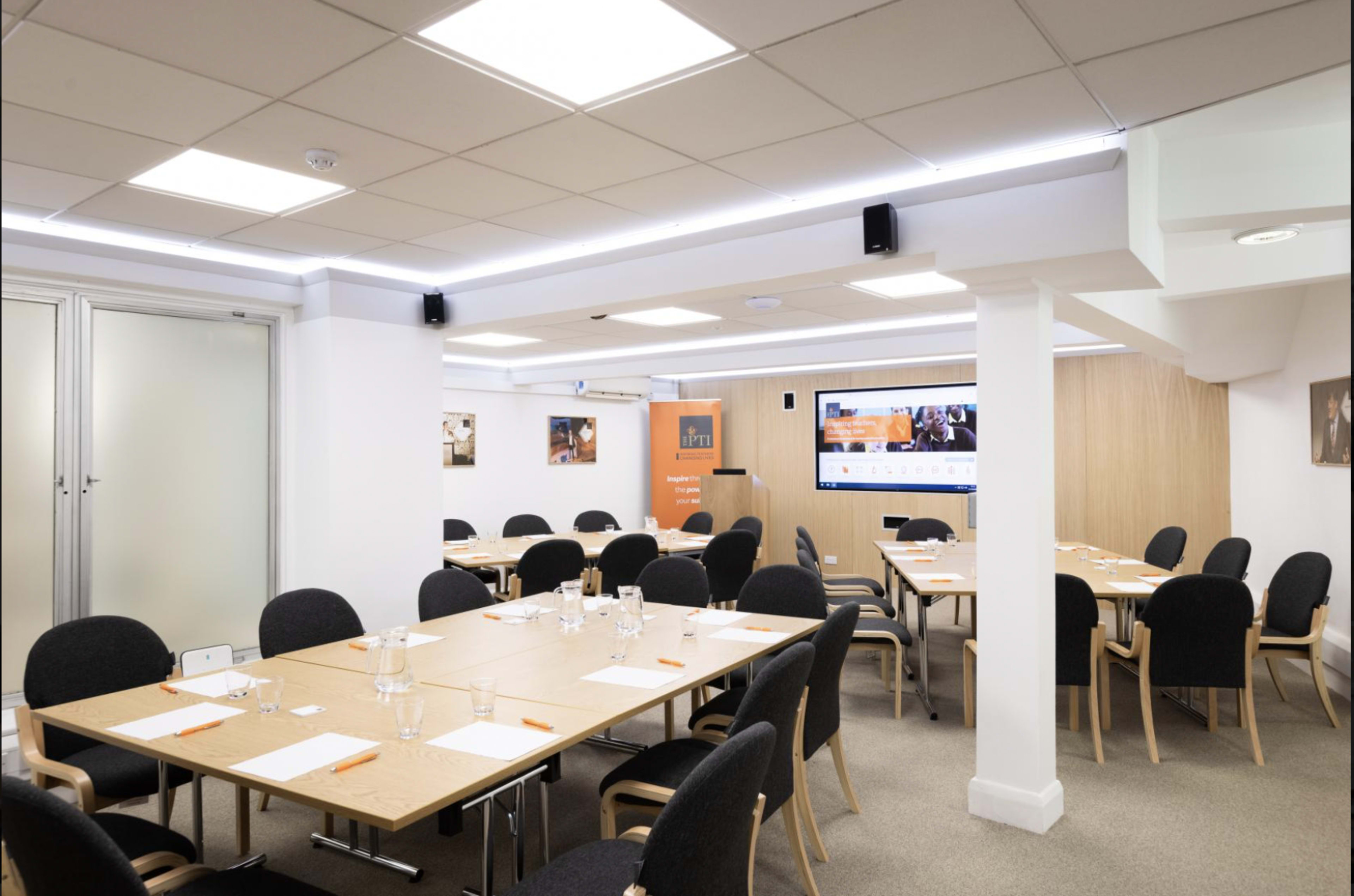 A modern conference room with a large wooden table surrounded by black chairs and a wall-mounted screen displaying a presentation.
