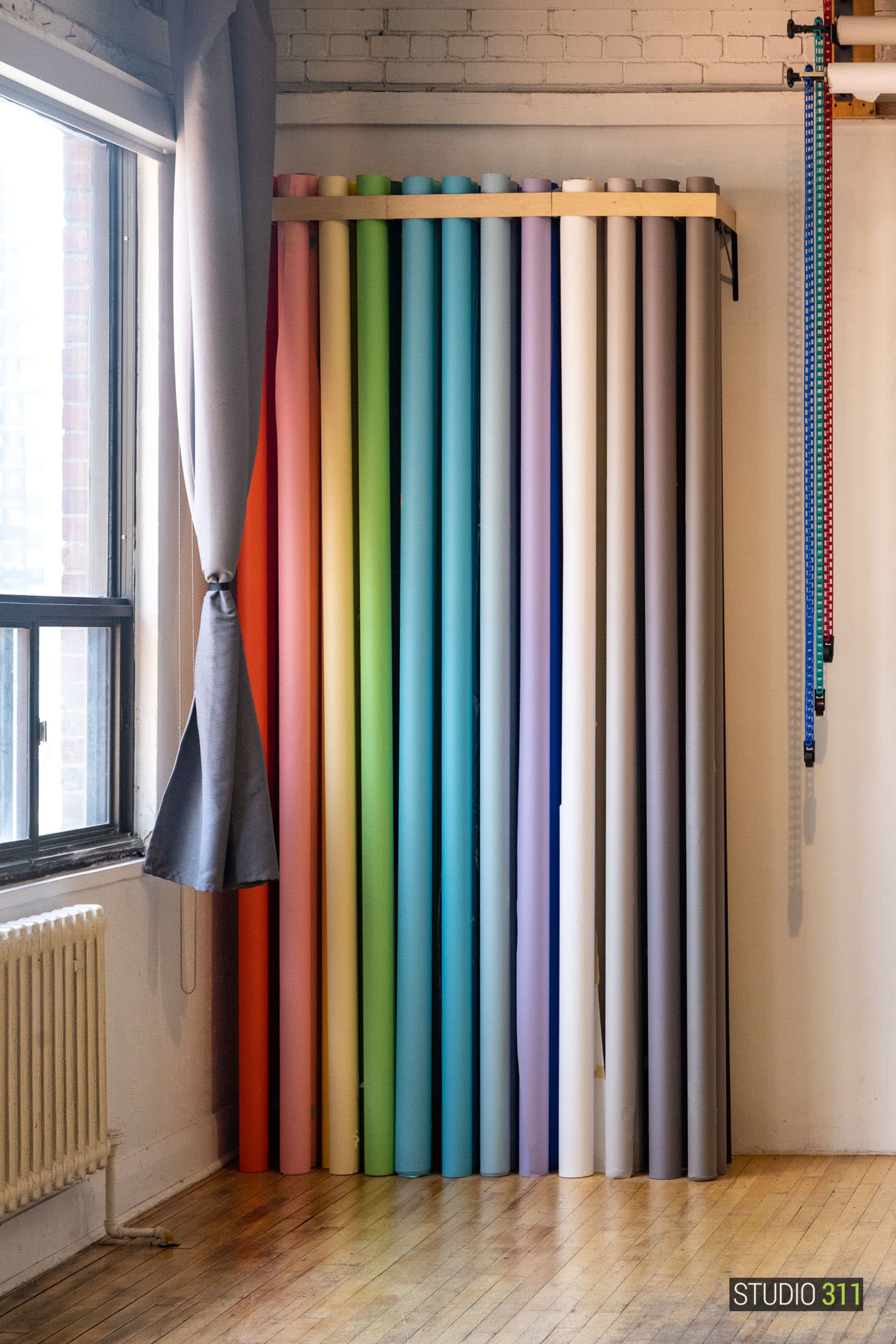 The image shows a collection of colorful paper rolls arranged vertically against a wall in a studio, with a window and curtain beside them.