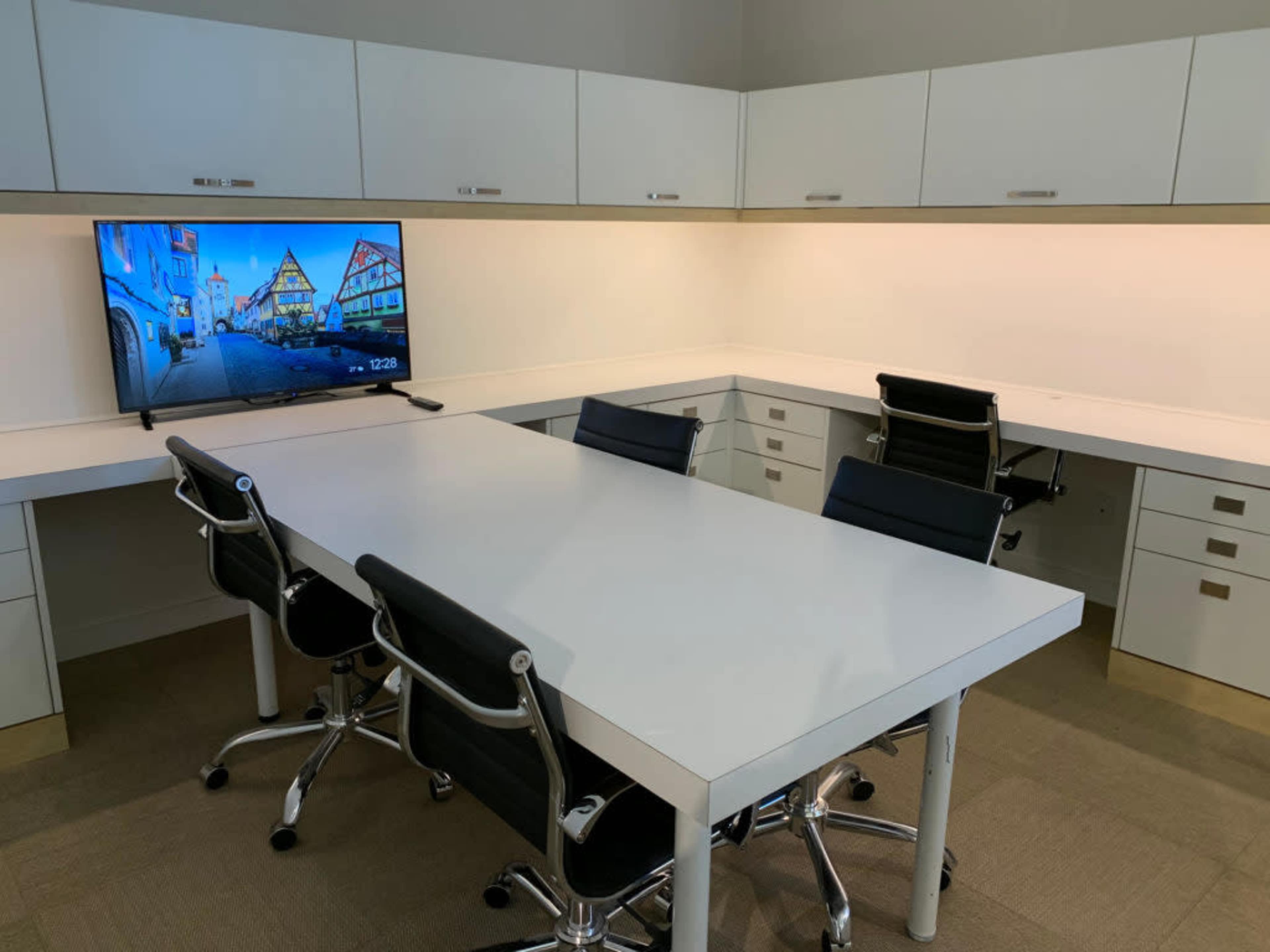 A modern conference room features a large white table surrounded by black chairs and a mounted television on the wall.