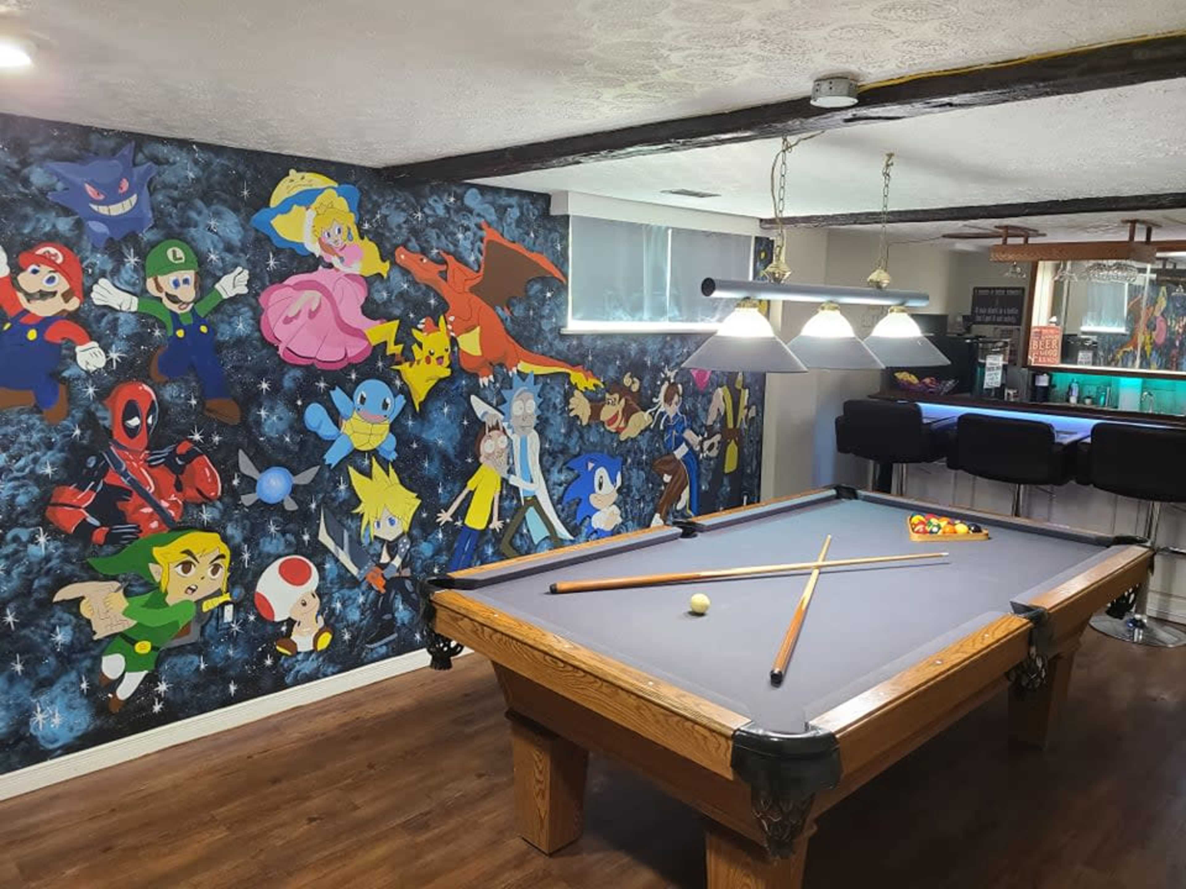 A pool table is centered in a room with a wall covered in colorful cartoon character murals, including superheroes and video game icons.