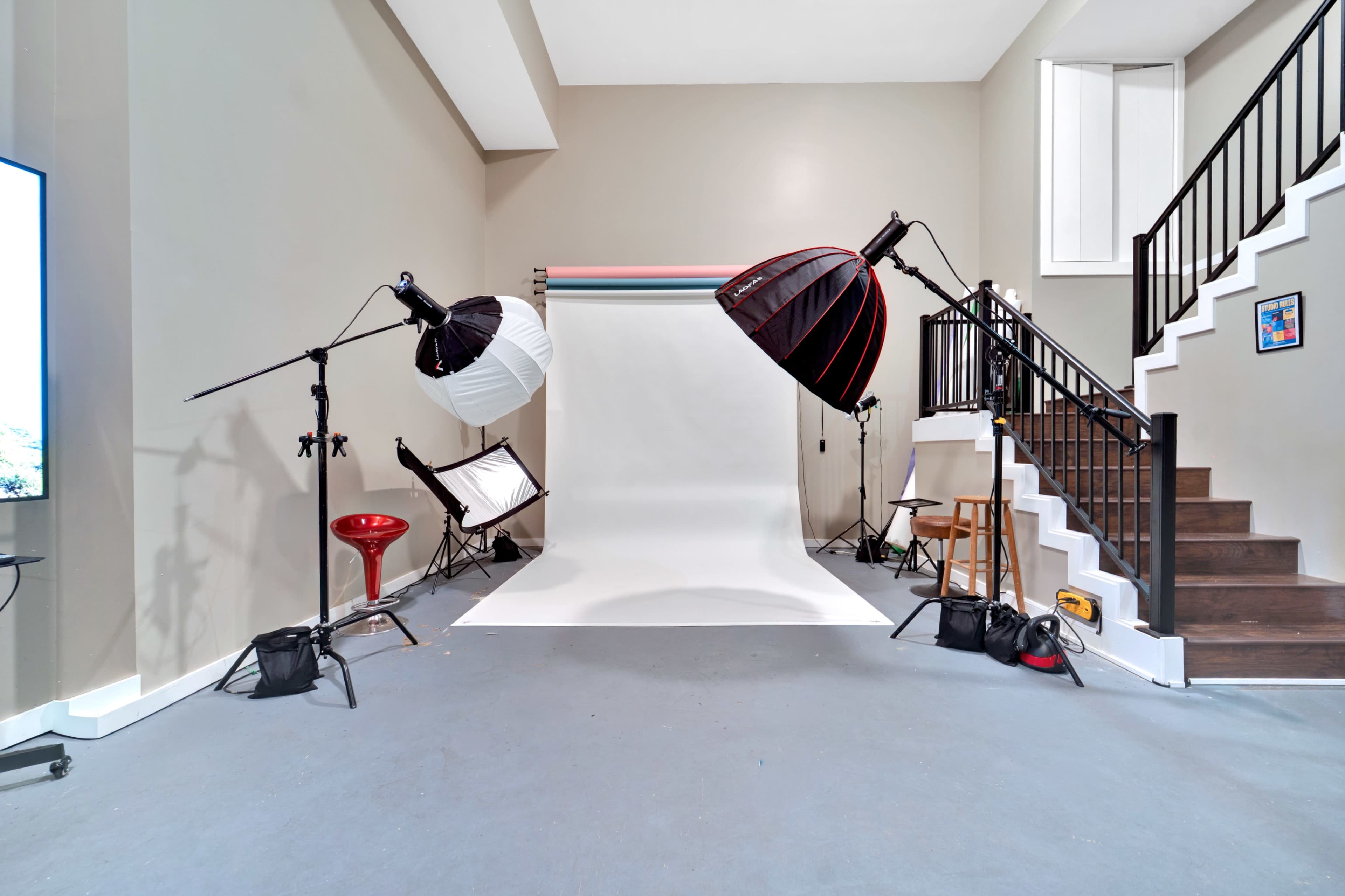 The image shows a photography studio with two large light setups, a white backdrop, and a staircase leading to an upper level.