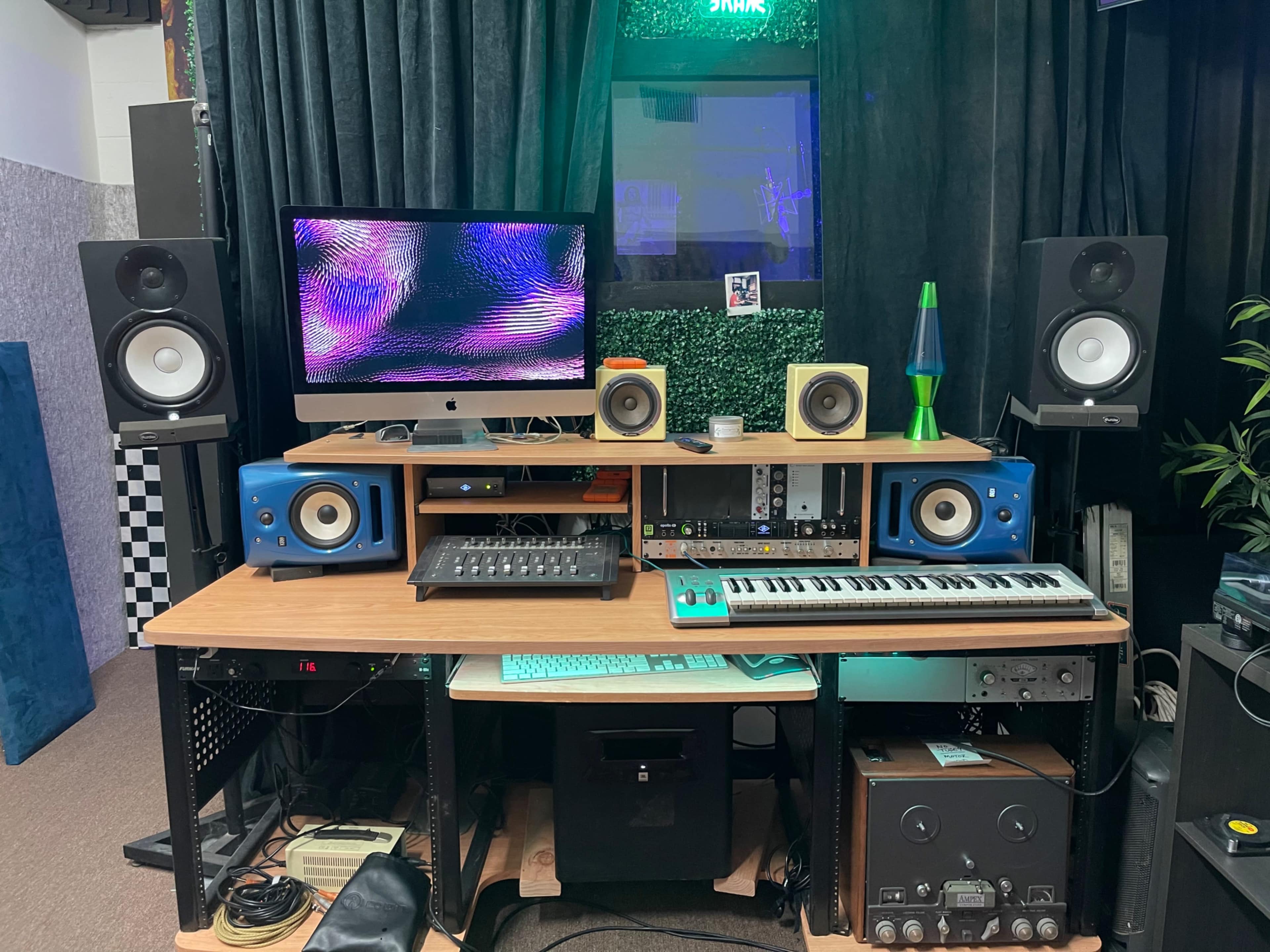A music production workstation features a computer, audio monitors, a MIDI keyboard, and various recording equipment arranged on a wooden desk.