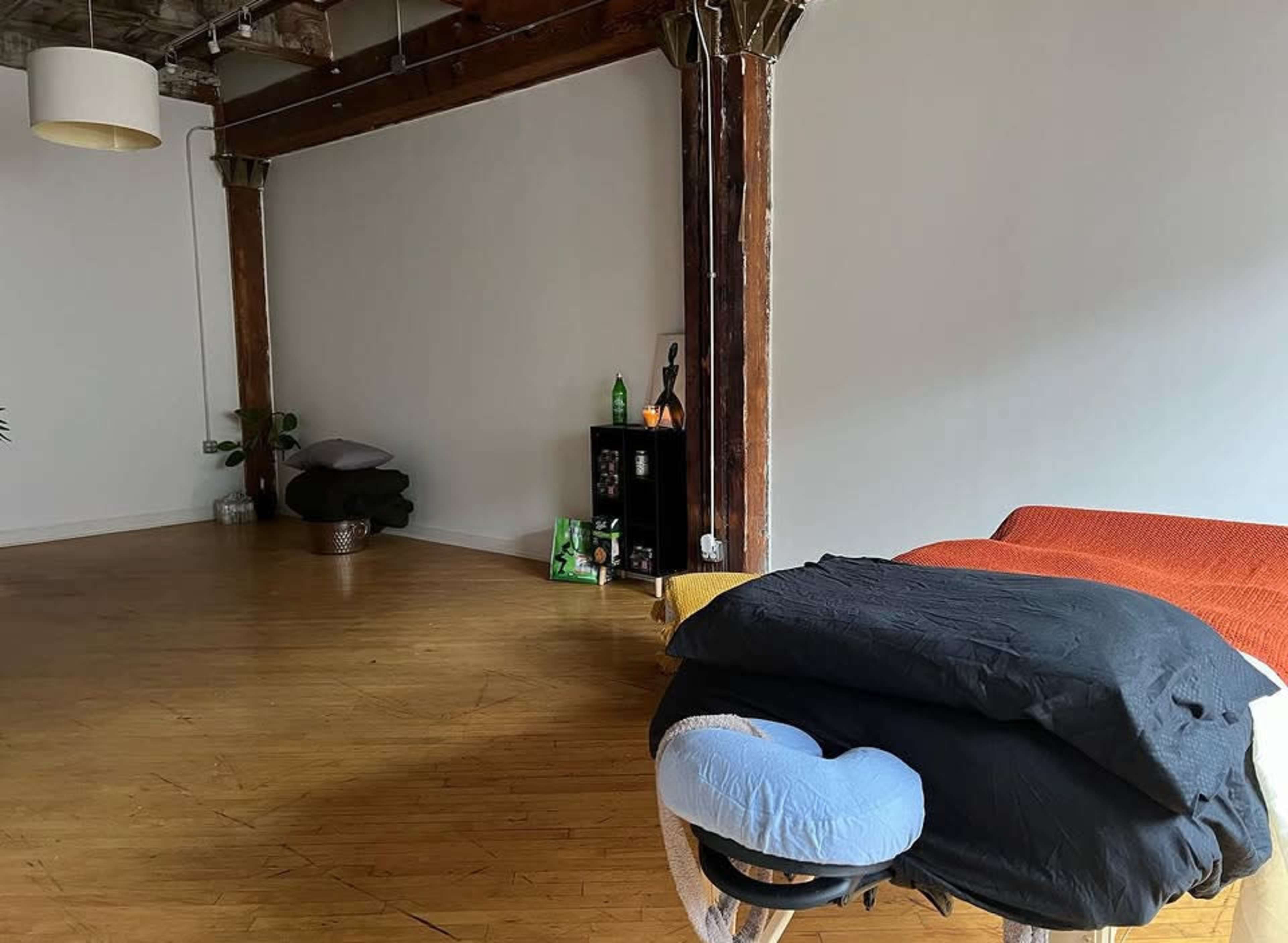 The image shows a spacious, empty room with wooden flooring, exposed beams, and a massage table covered with various colored blankets, along with a small shelf displaying a few items against a plain wall.