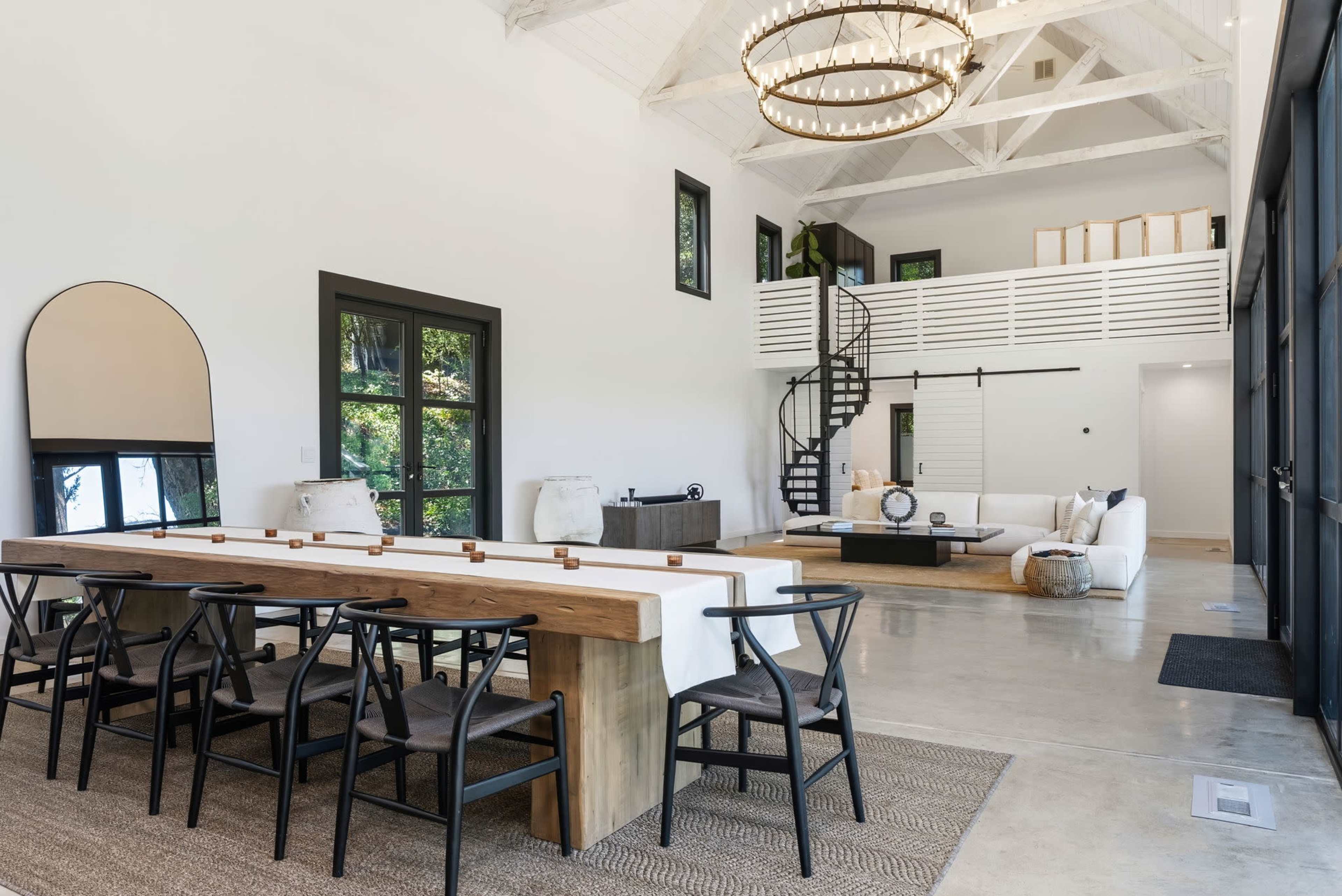 The image shows a modern interior of a spacious living area with a large wooden dining table, black chairs, and a spiral staircase leading to a lofted space.