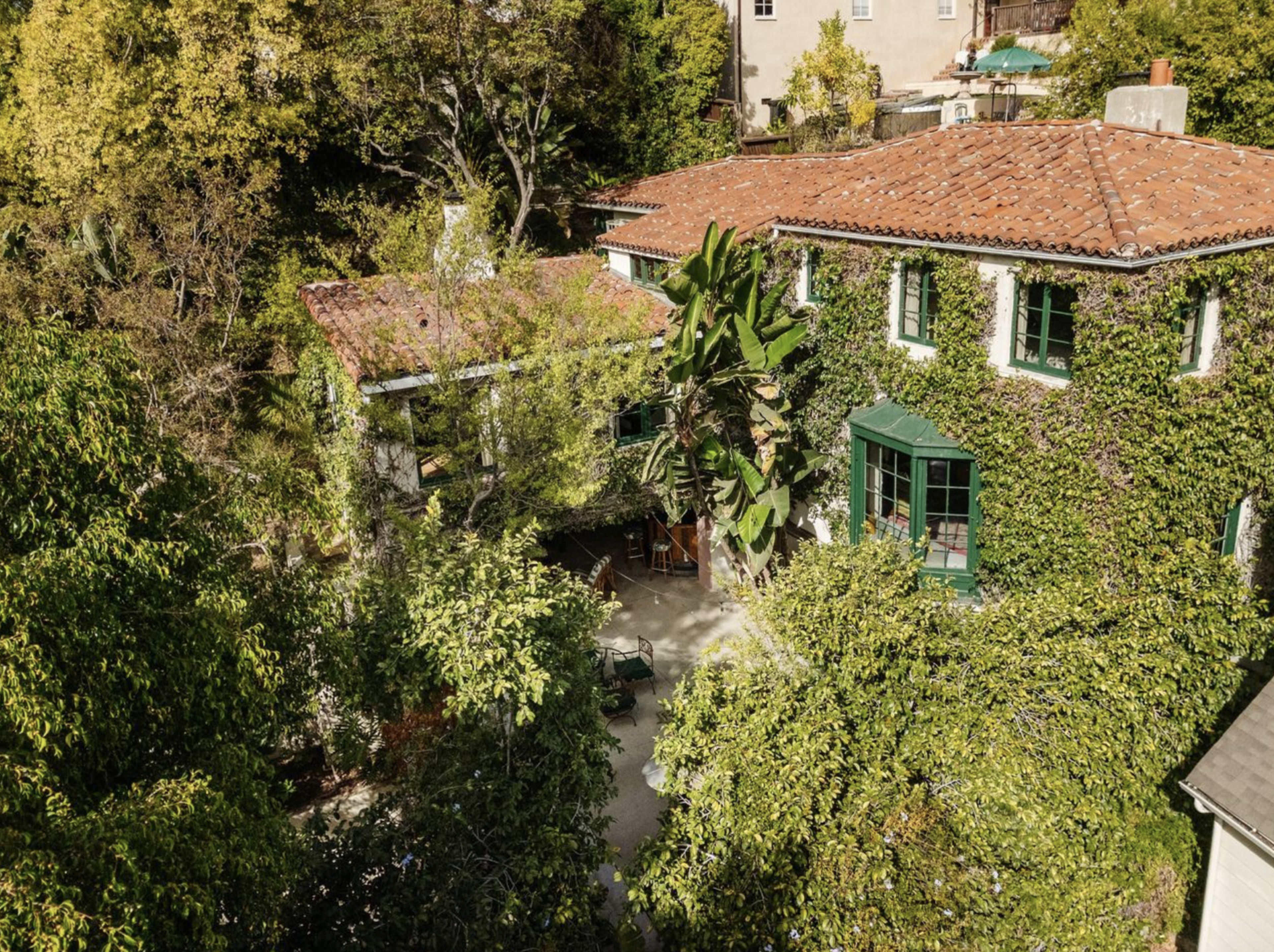 A two-story house covered in green ivy features a terracotta tile roof and surrounded by trees and foliage.