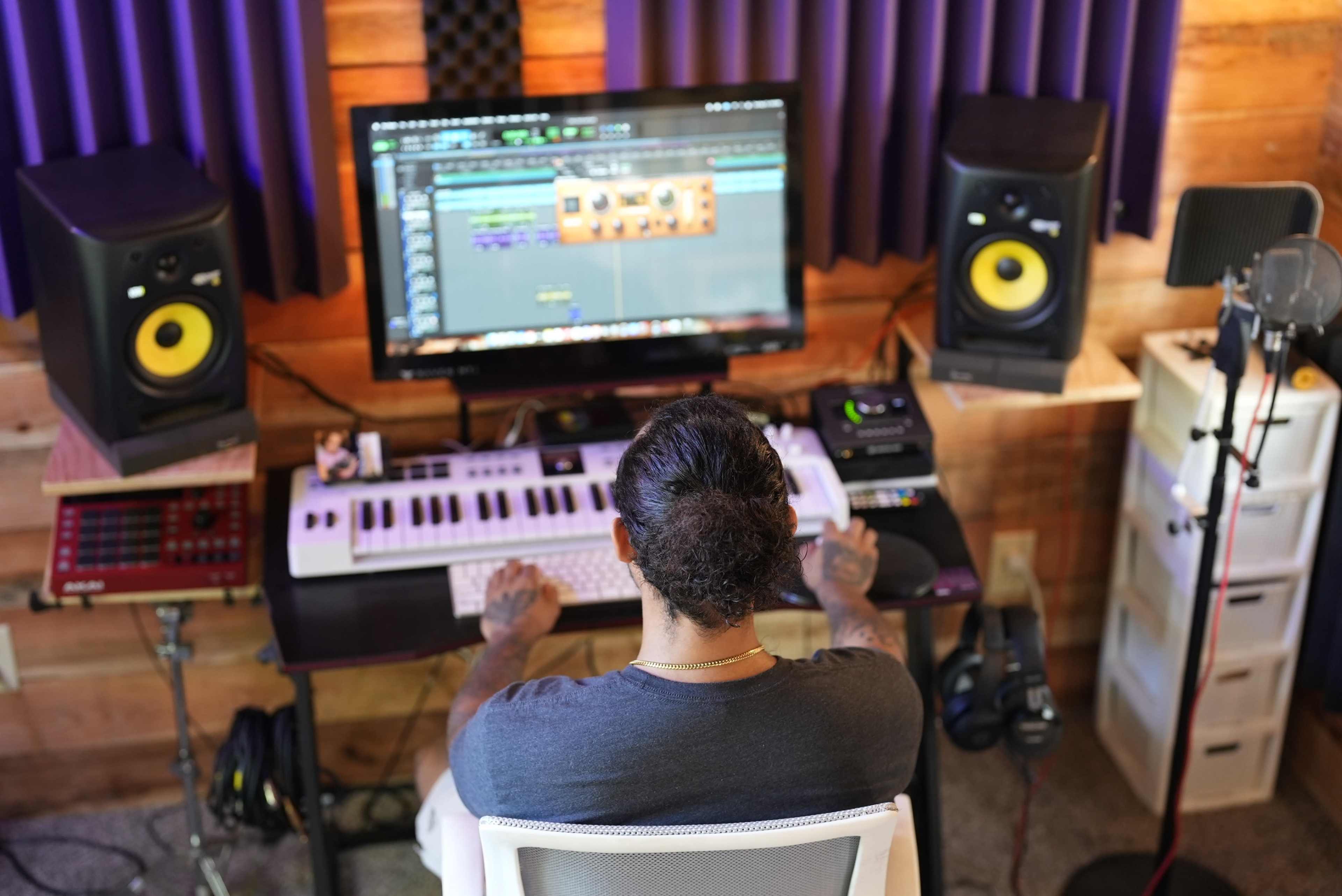 A person with a ponytail sits at a keyboard in front of a computer screen, surrounded by studio monitors and recording equipment in a recording studio.