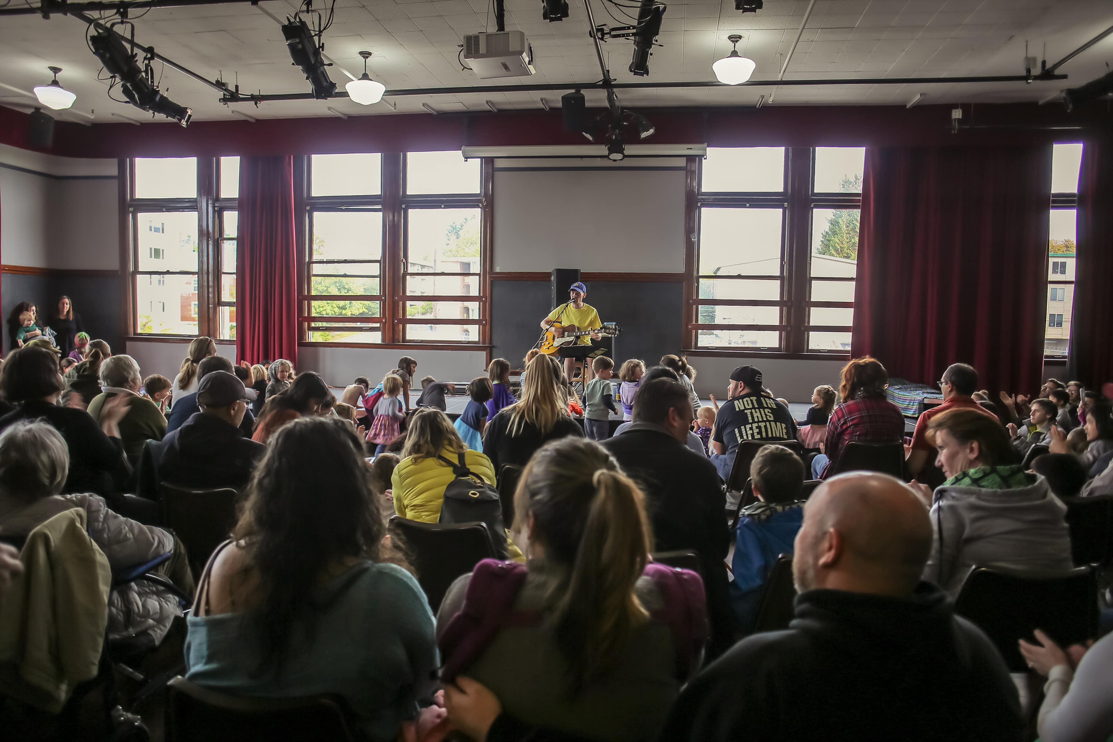 A performer plays guitar on a small stage in front of an audience seated in a room with large windows.