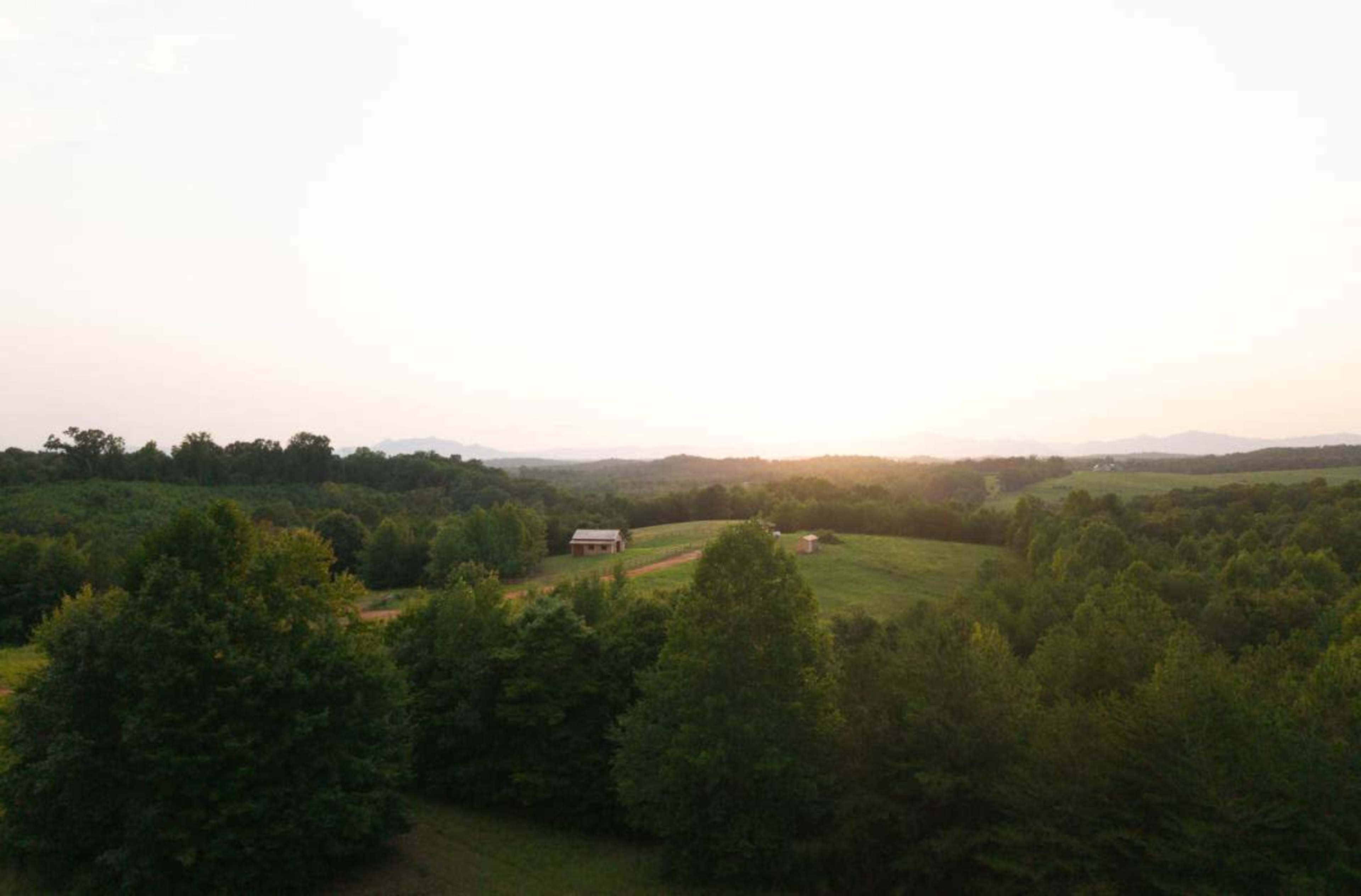 A rural landscape features rolling hills, lush green trees, and a distant sunset illuminating a small wooden barn.