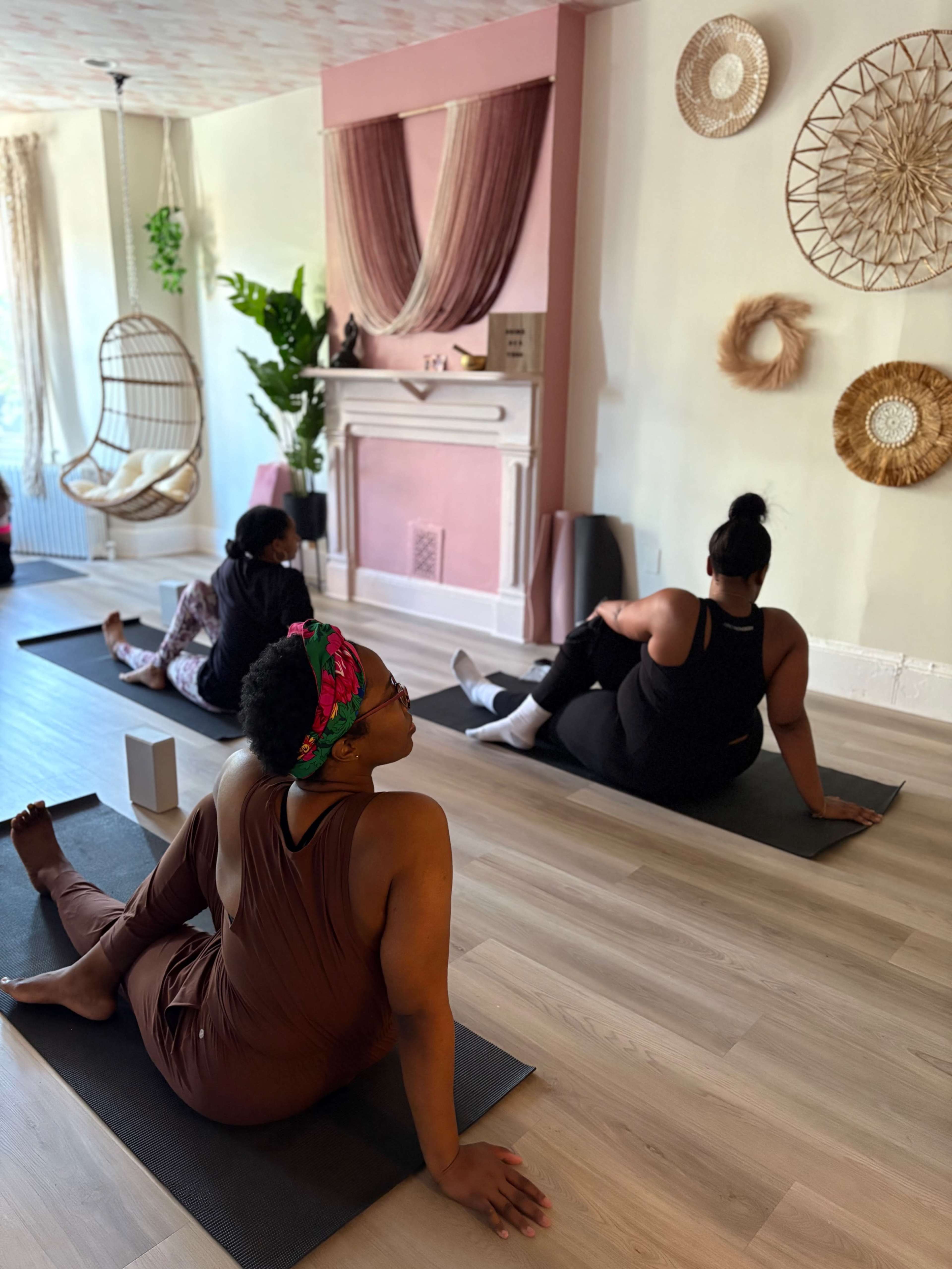 Three people are practicing yoga on mats in a brightly lit room with decorative wall hangings and plants.