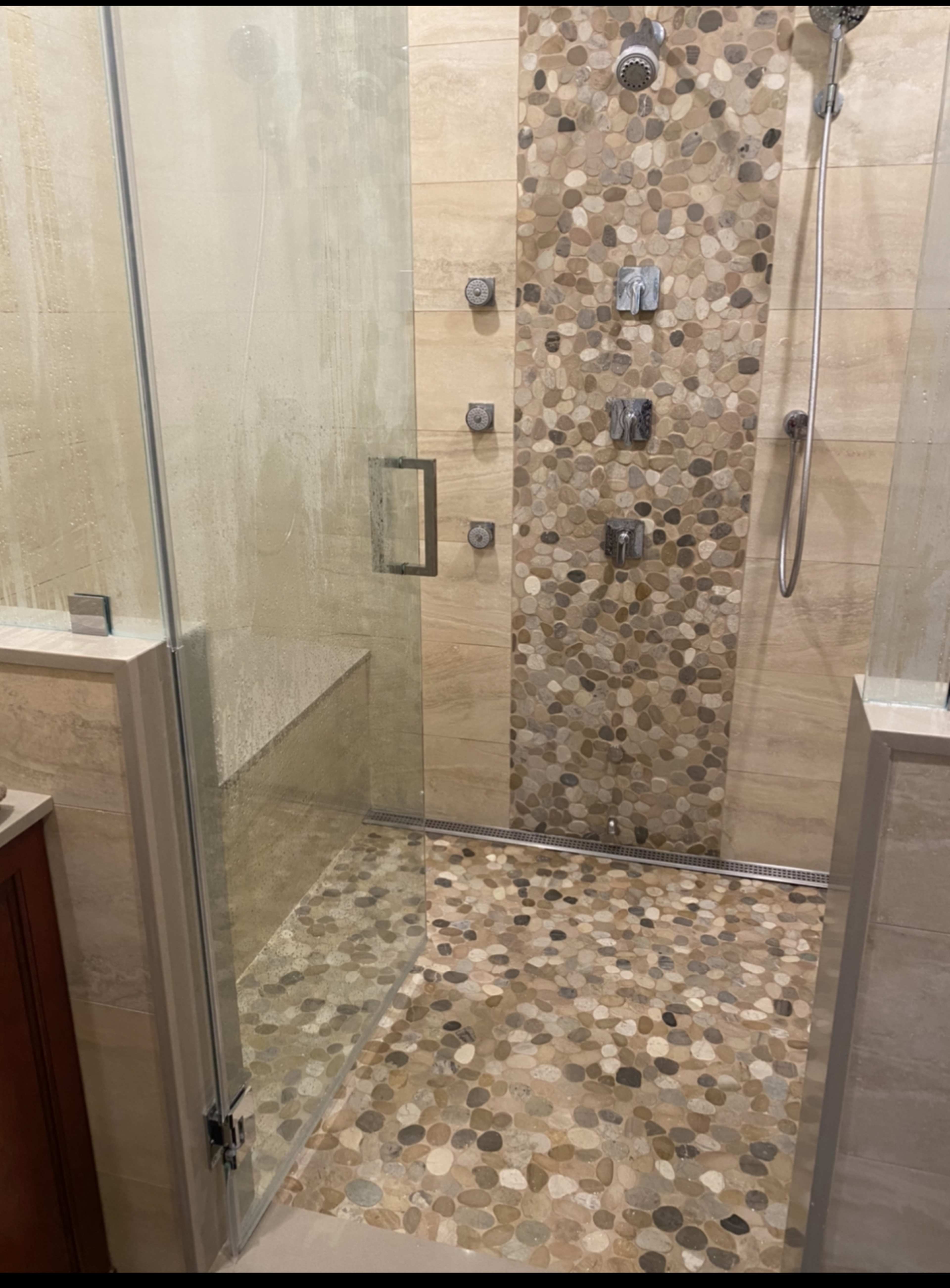 A modern shower area with a glass enclosure, featuring a stone mosaic floor and a tiled wall with multiple shower fixtures.