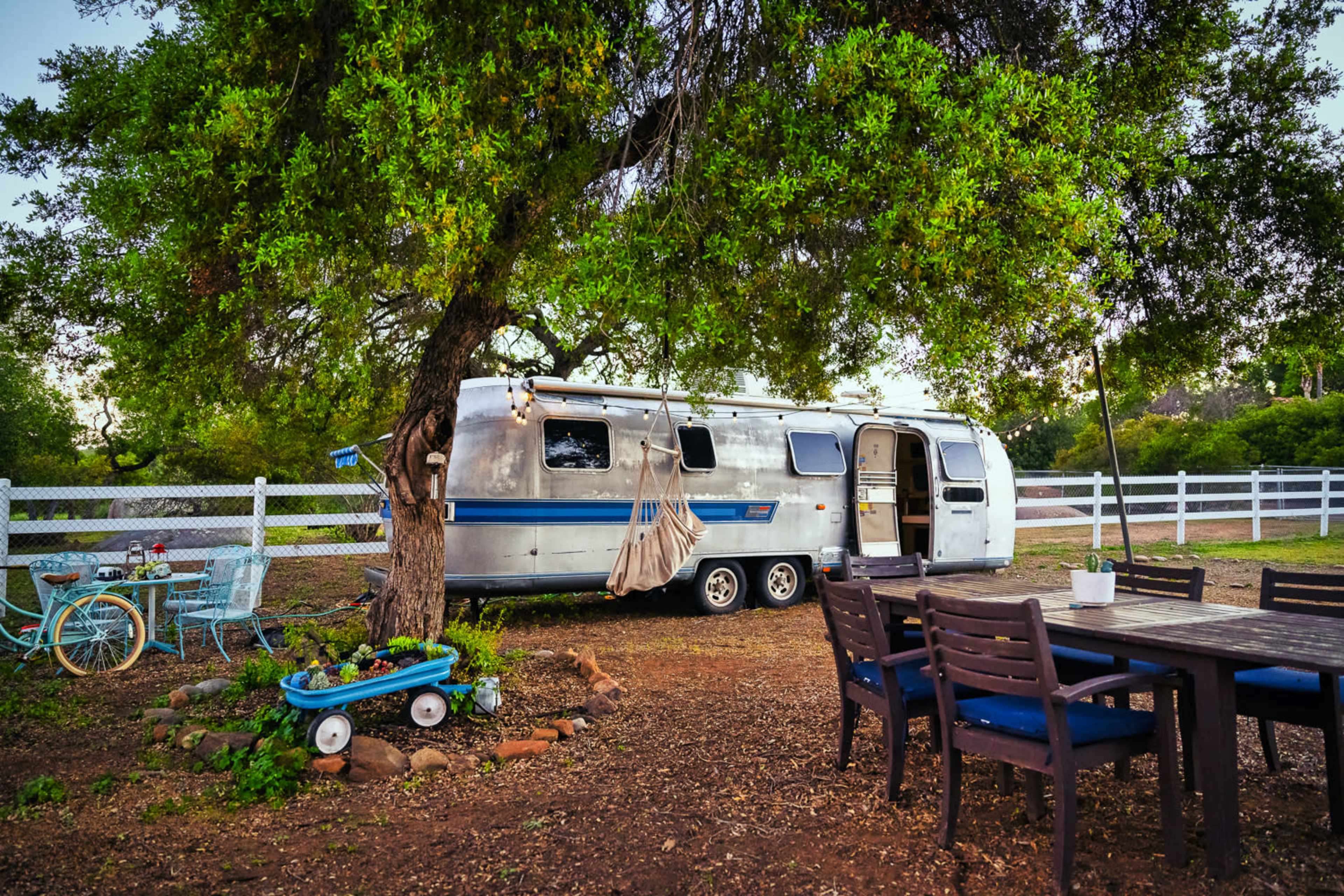 A silver Airstream trailer is parked under a large tree next to a wooden dining table and chairs in a grassy outdoor area.