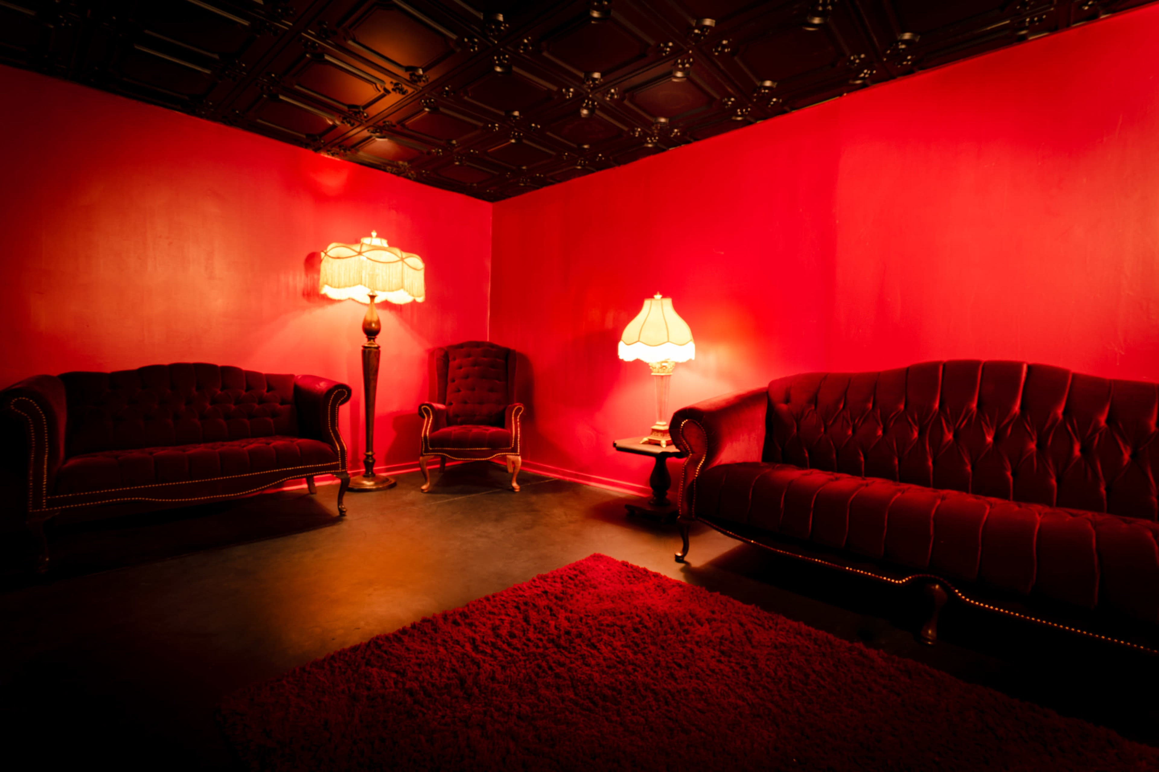 The image shows a room with red walls, two ornate sofas, a chair, and two lamps providing soft lighting.