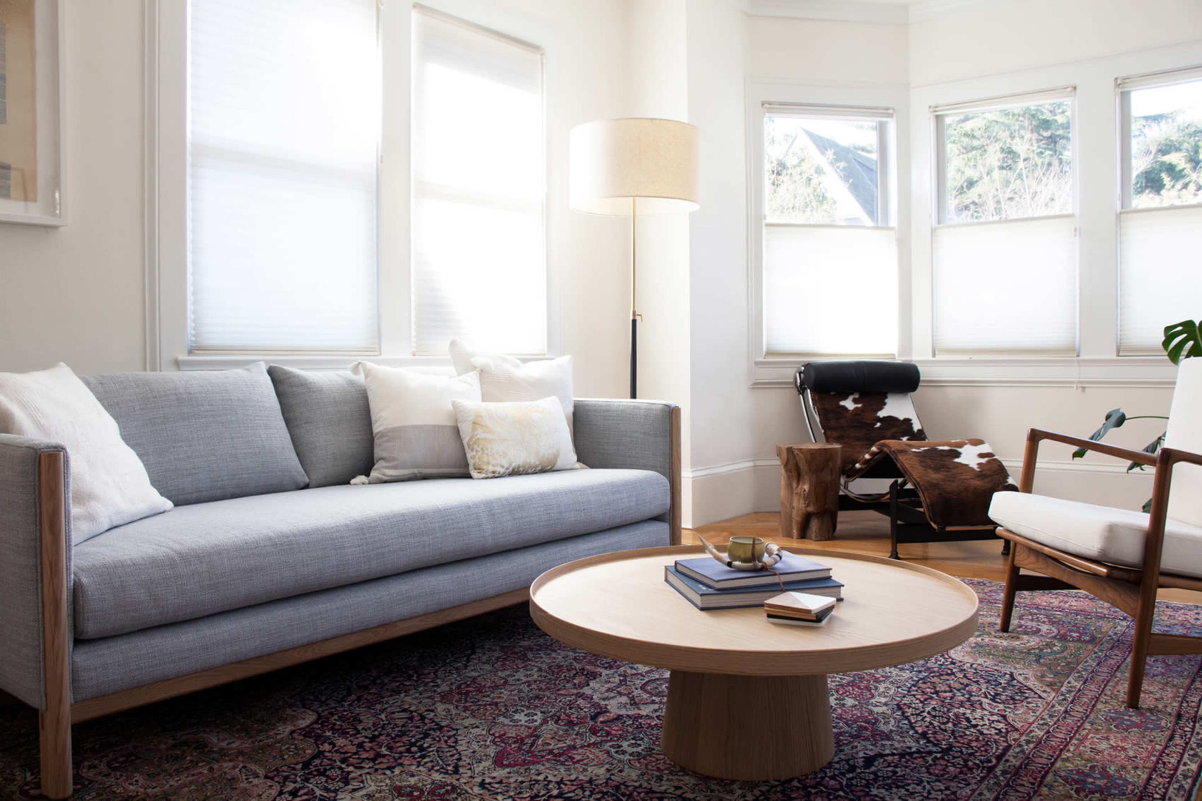 A modern living room with a gray sofa, a round wooden coffee table, and various pieces of furniture, illuminated by natural light from several windows.