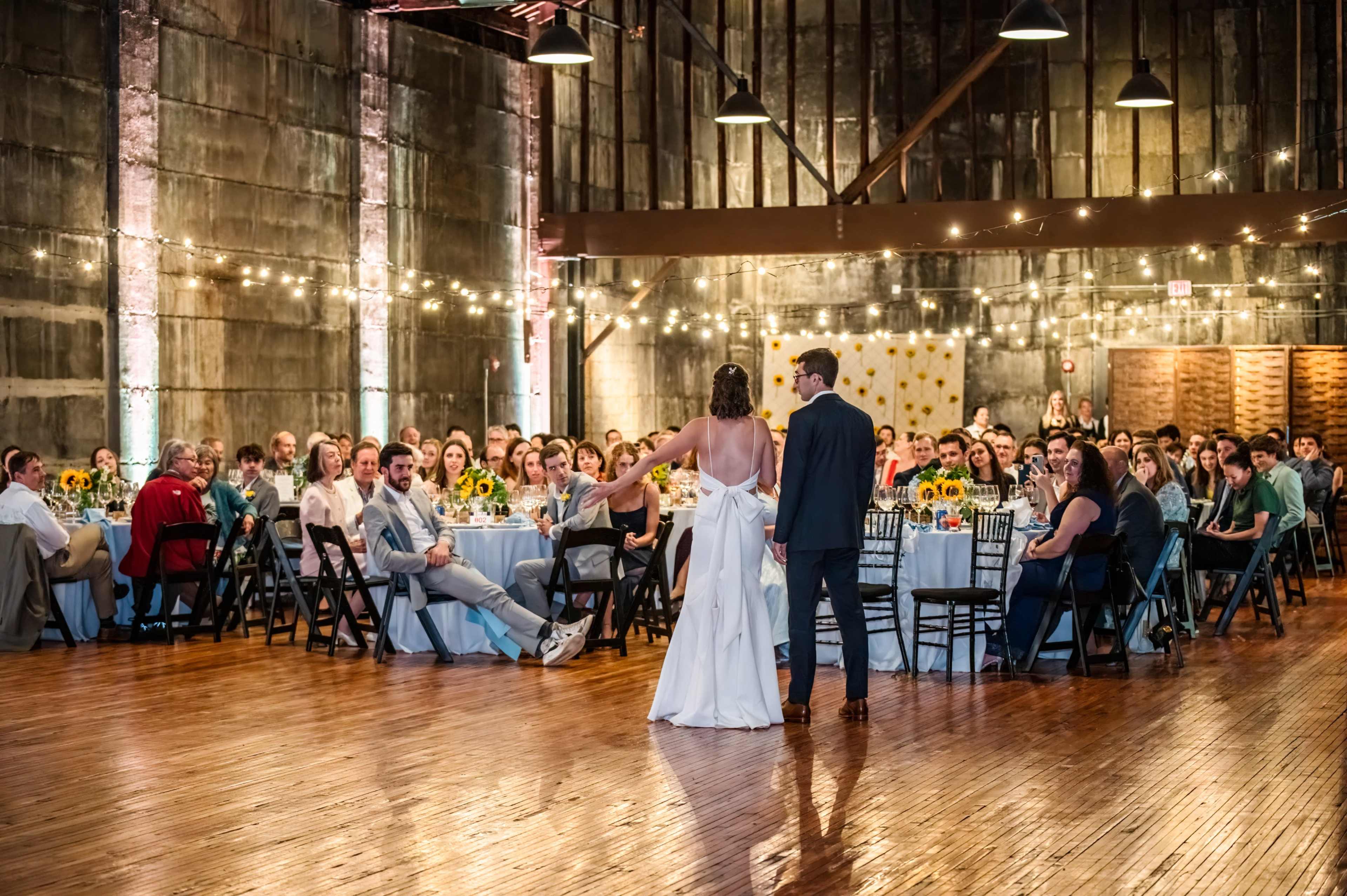 A couple stands together at the front of a rustic venue, addressing an audience seated at tables adorned with sunflower centerpieces.
