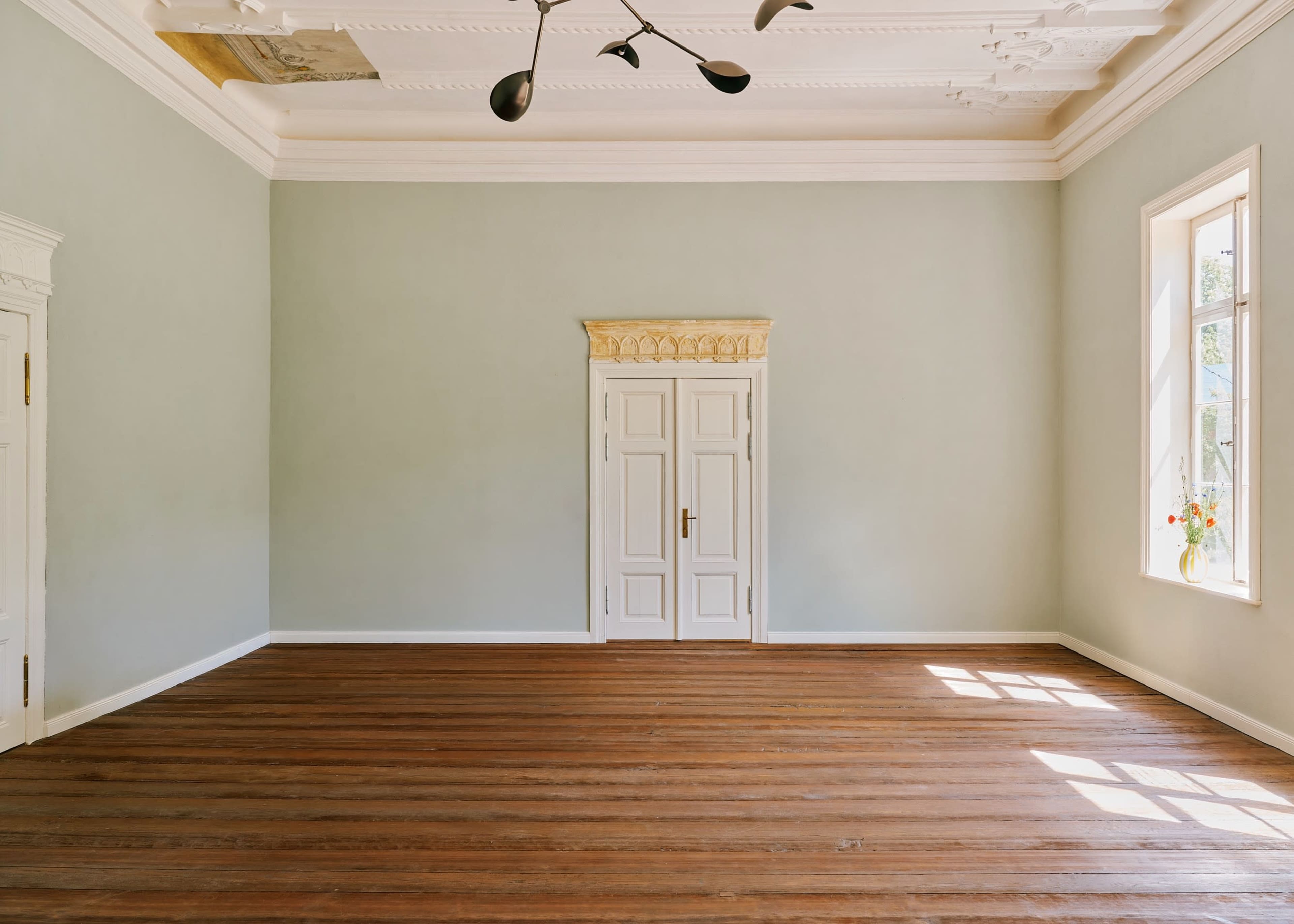 The image shows a spacious room with light green walls, a single window allowing natural light, and polished wooden flooring.