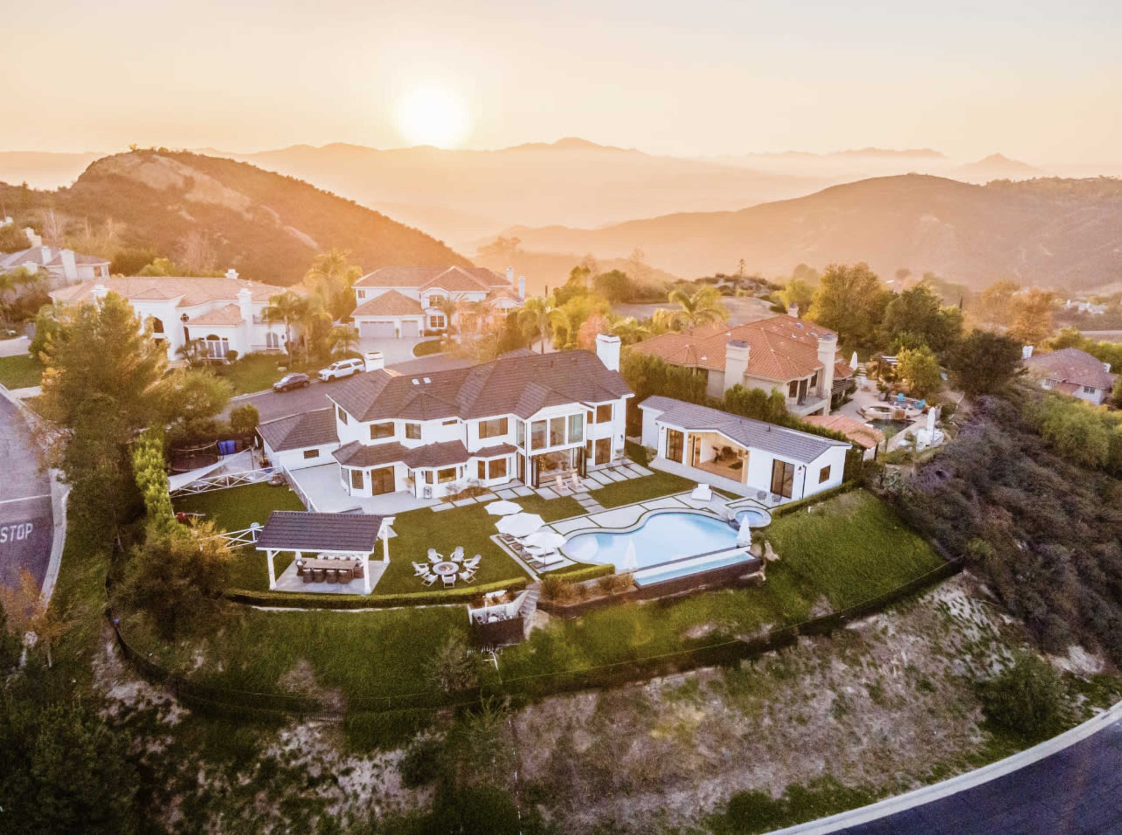A large modern house with a pool is situated on a hillside overlooking a sunset and surrounding mountains.