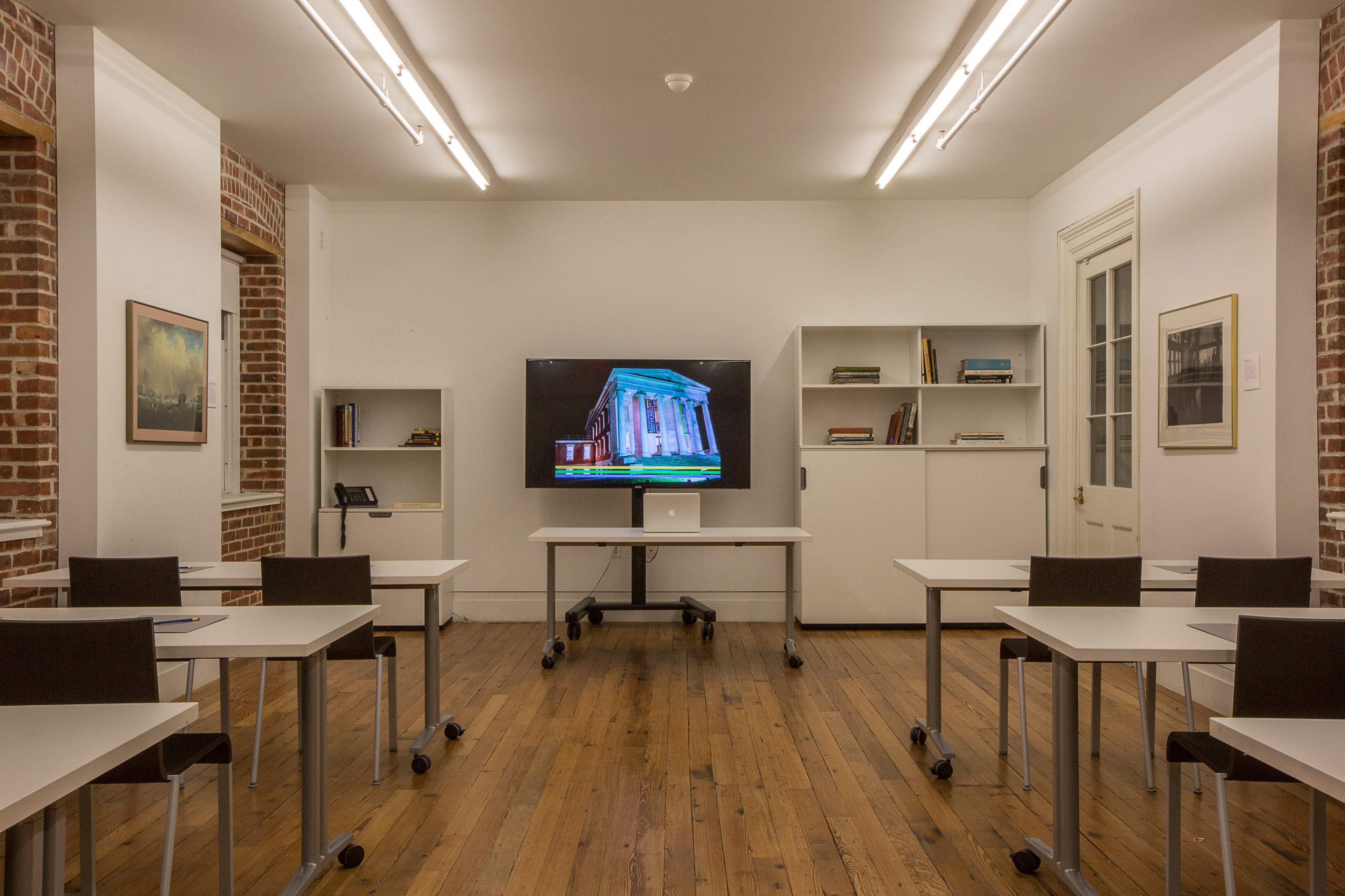 A classroom is arranged with several tables and chairs facing a large television mounted on the wall, surrounded by bookshelves against exposed brick walls.