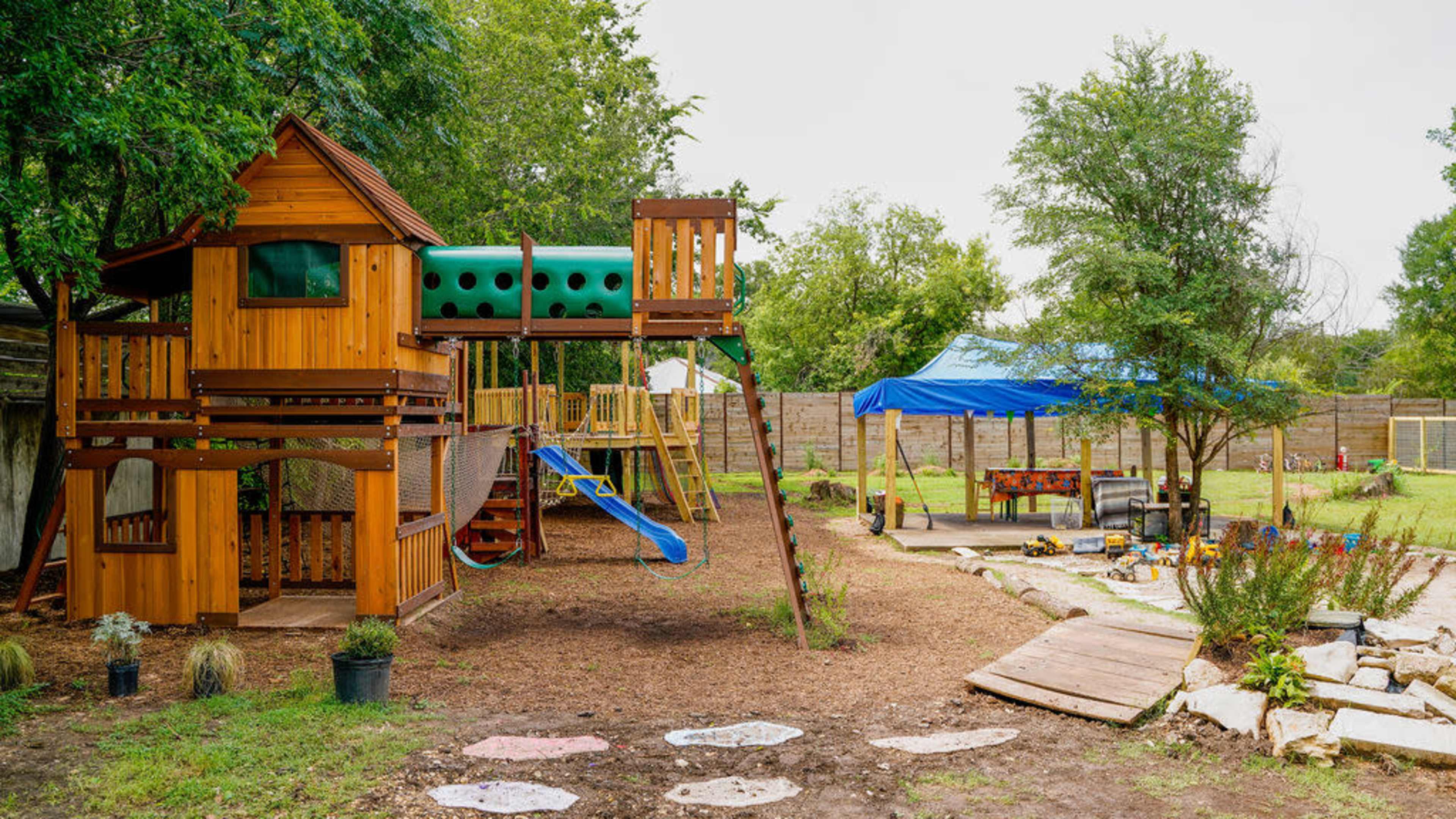 The image shows a wooden playset with a slide and climbing area alongside a blue tent and a garden area with various plants and toys.