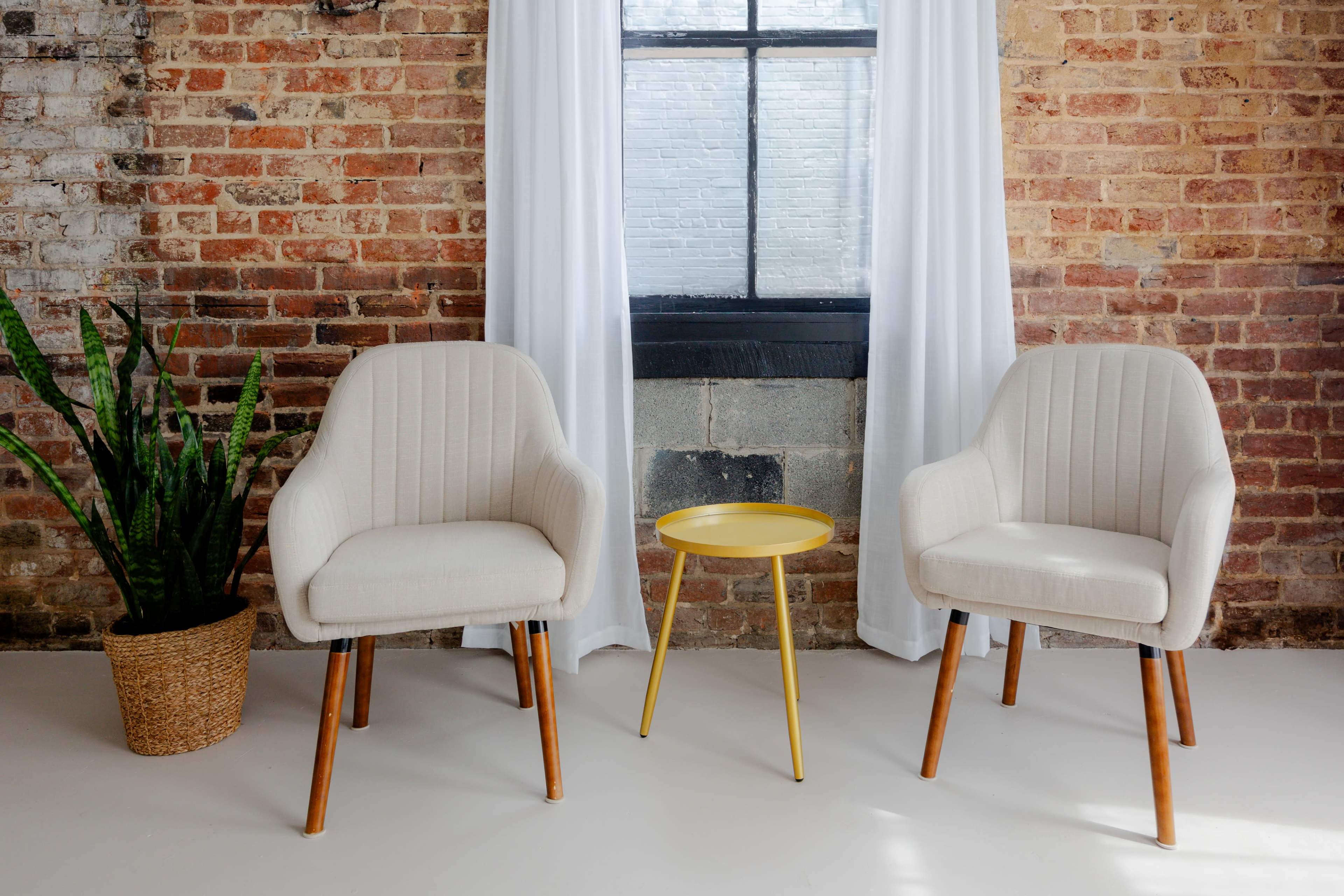 Two light-colored chairs with wooden legs placed beside a small yellow table, against a backdrop of a brick wall and a large window with white curtains.