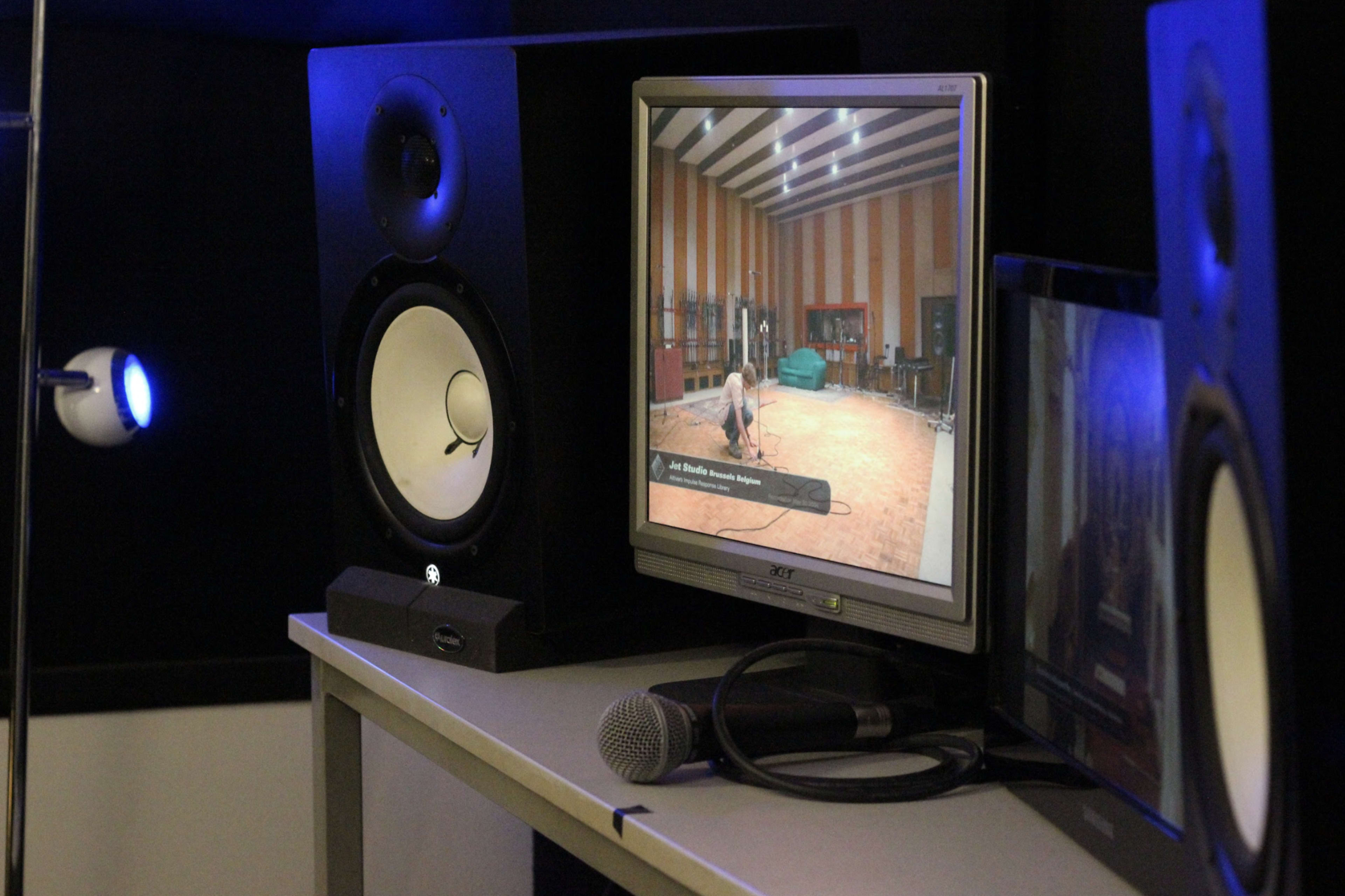 A computer monitor displays a video of a person in a recording studio, with audio speakers and a microphone nearby.