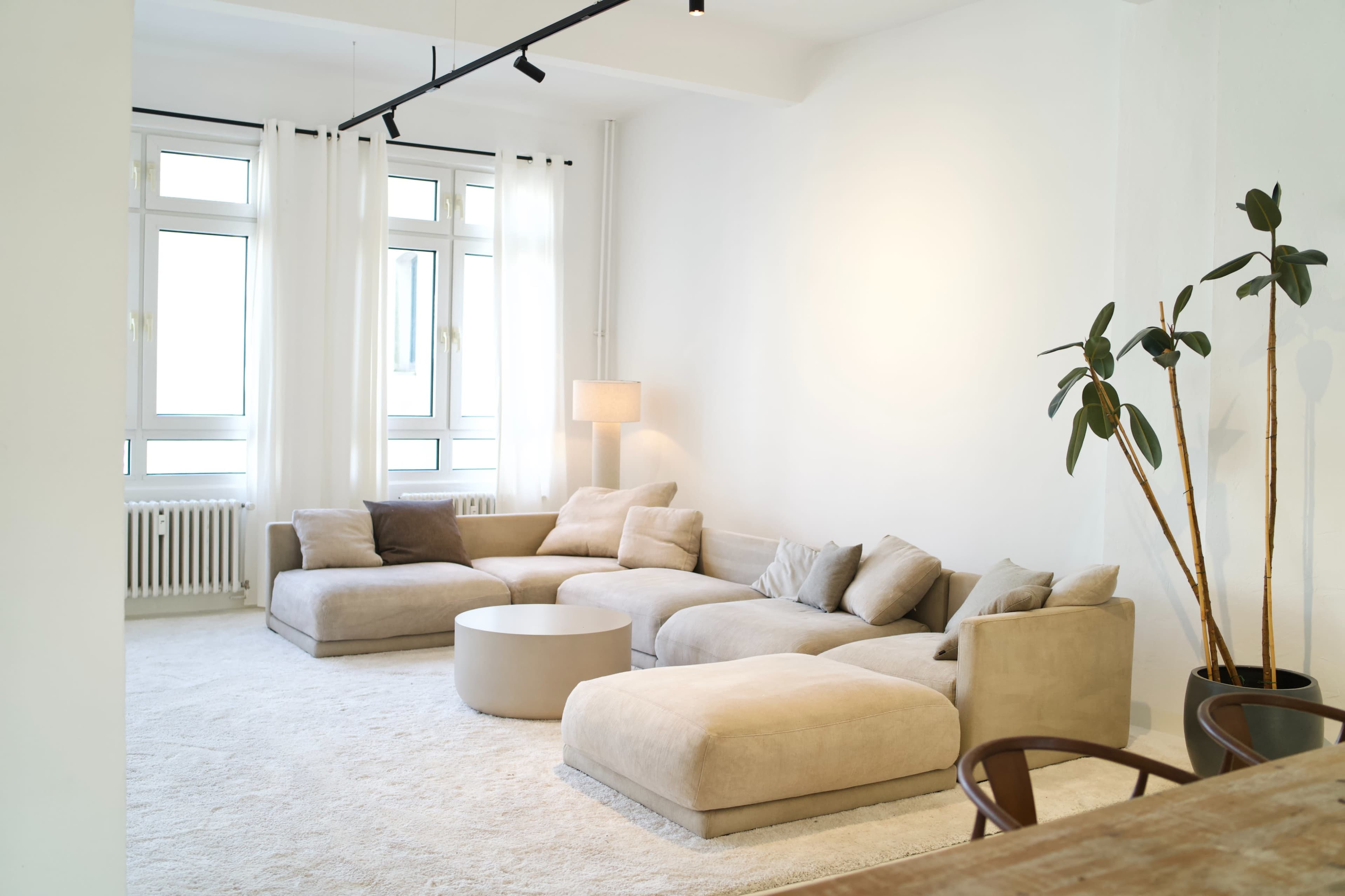 The image shows a modern living room with a large light-colored sectional sofa, a circular coffee table, and a potted plant, all set against white walls and large windows.