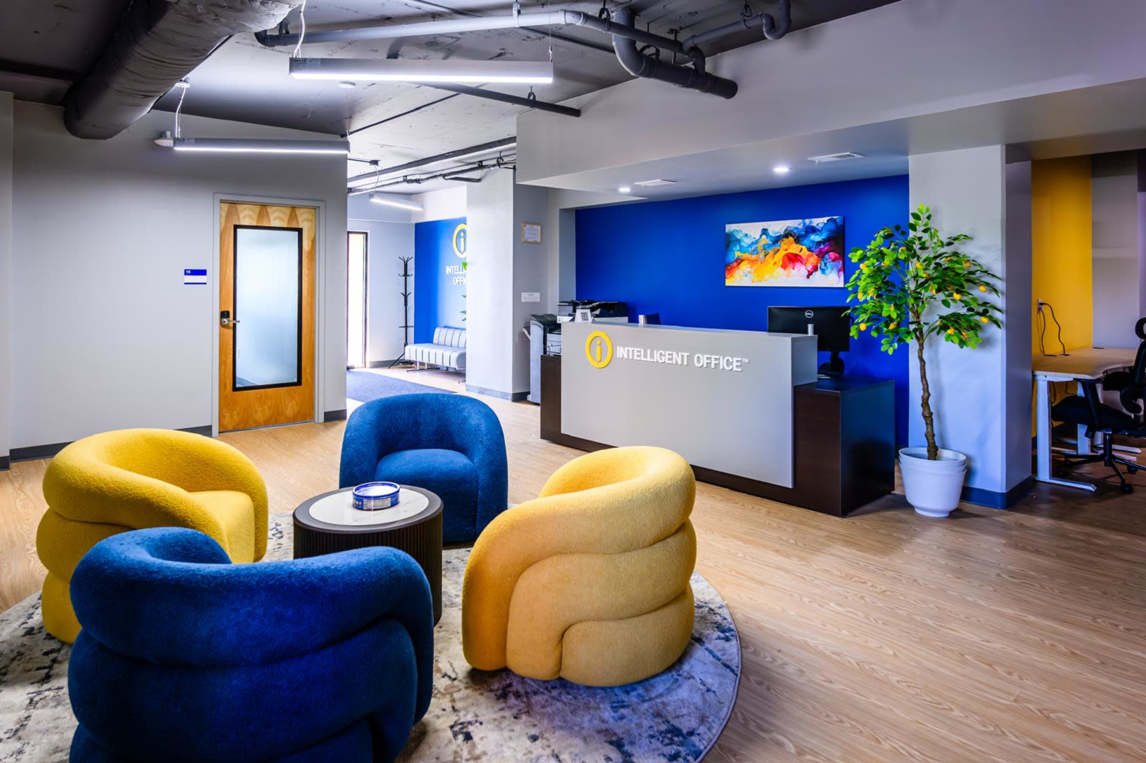 The image depicts a modern office reception area featuring a circular seating arrangement with blue and yellow chairs, a reception desk, and a colorful wall art display.