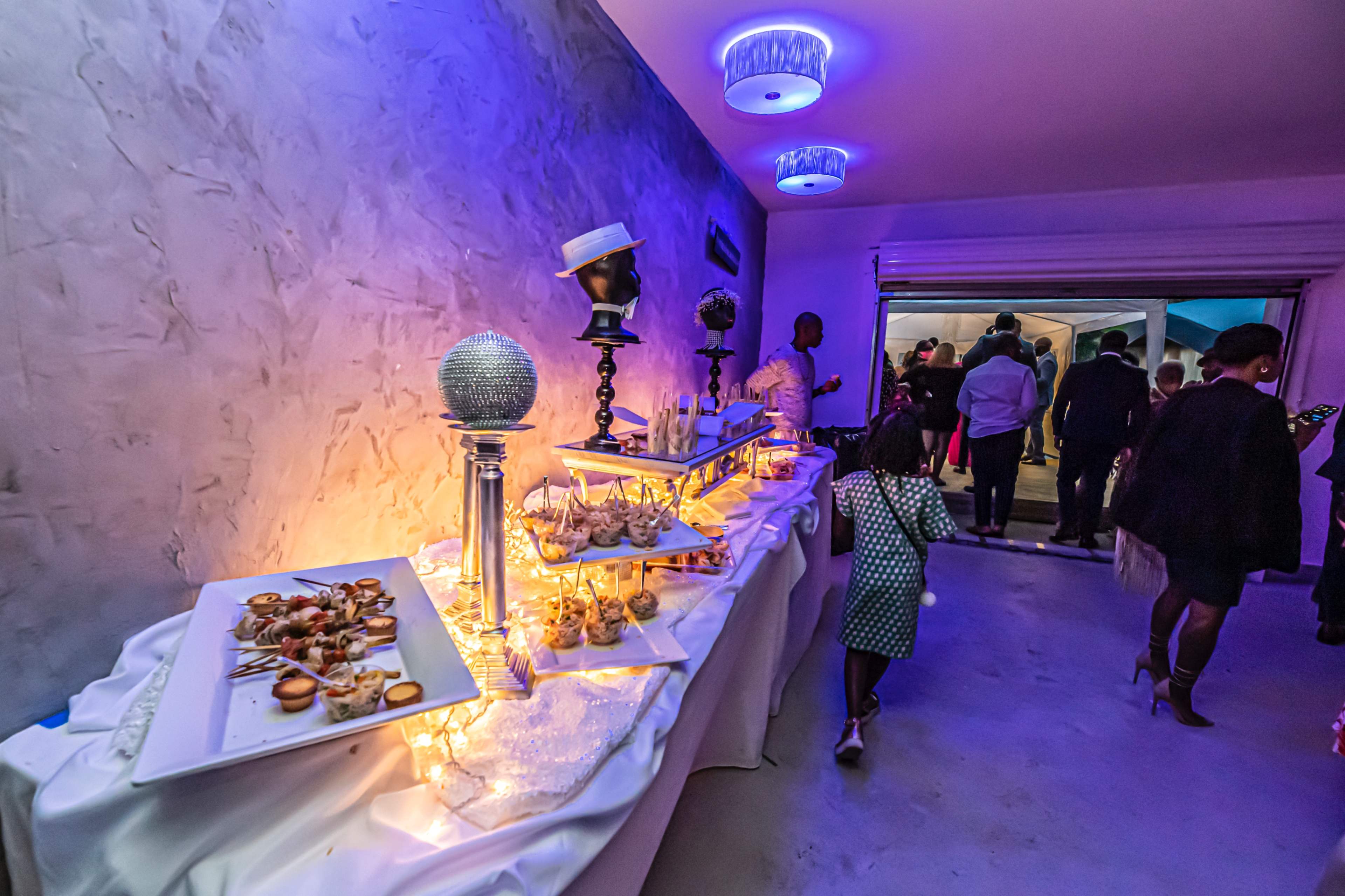 A buffet table adorned with various appetizers and decorative elements is set up in a dimly lit room, with guests mingling in the background.