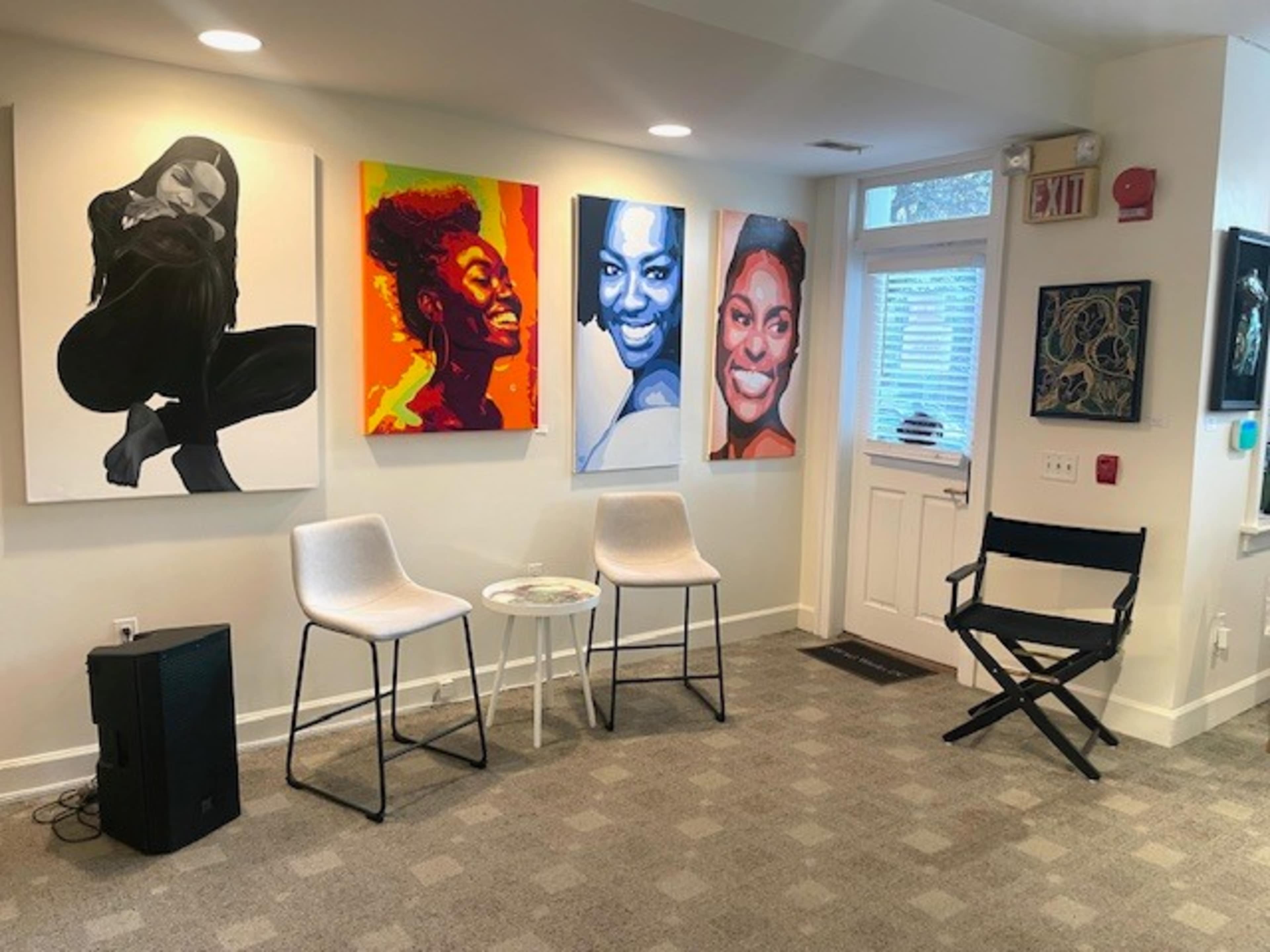 The image shows a modern gallery space featuring three colorful portraits on the walls, two white chairs and a small round table in front of them, and a director's chair nearby.