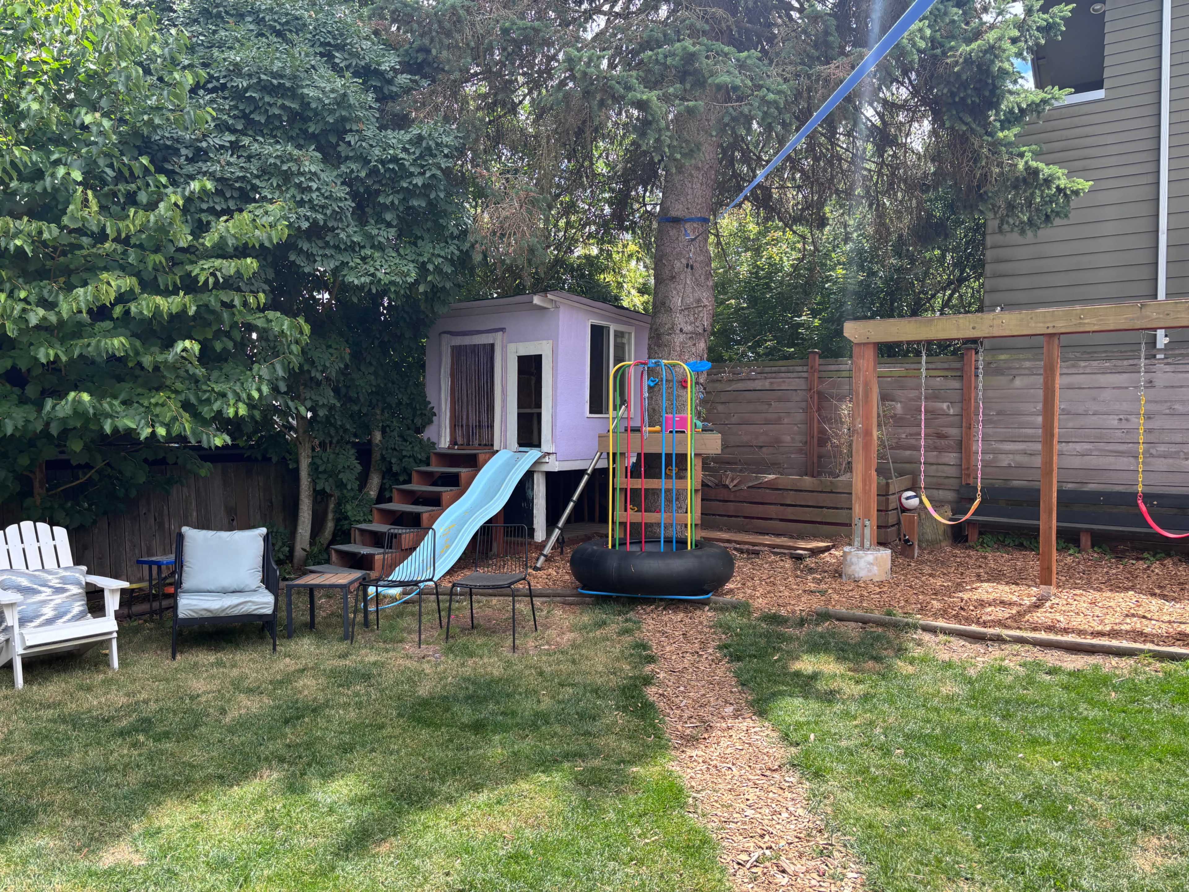 The image shows a backyard playground area featuring a playset with a slide, a treehouse, and swings, surrounded by grass and garden beds.