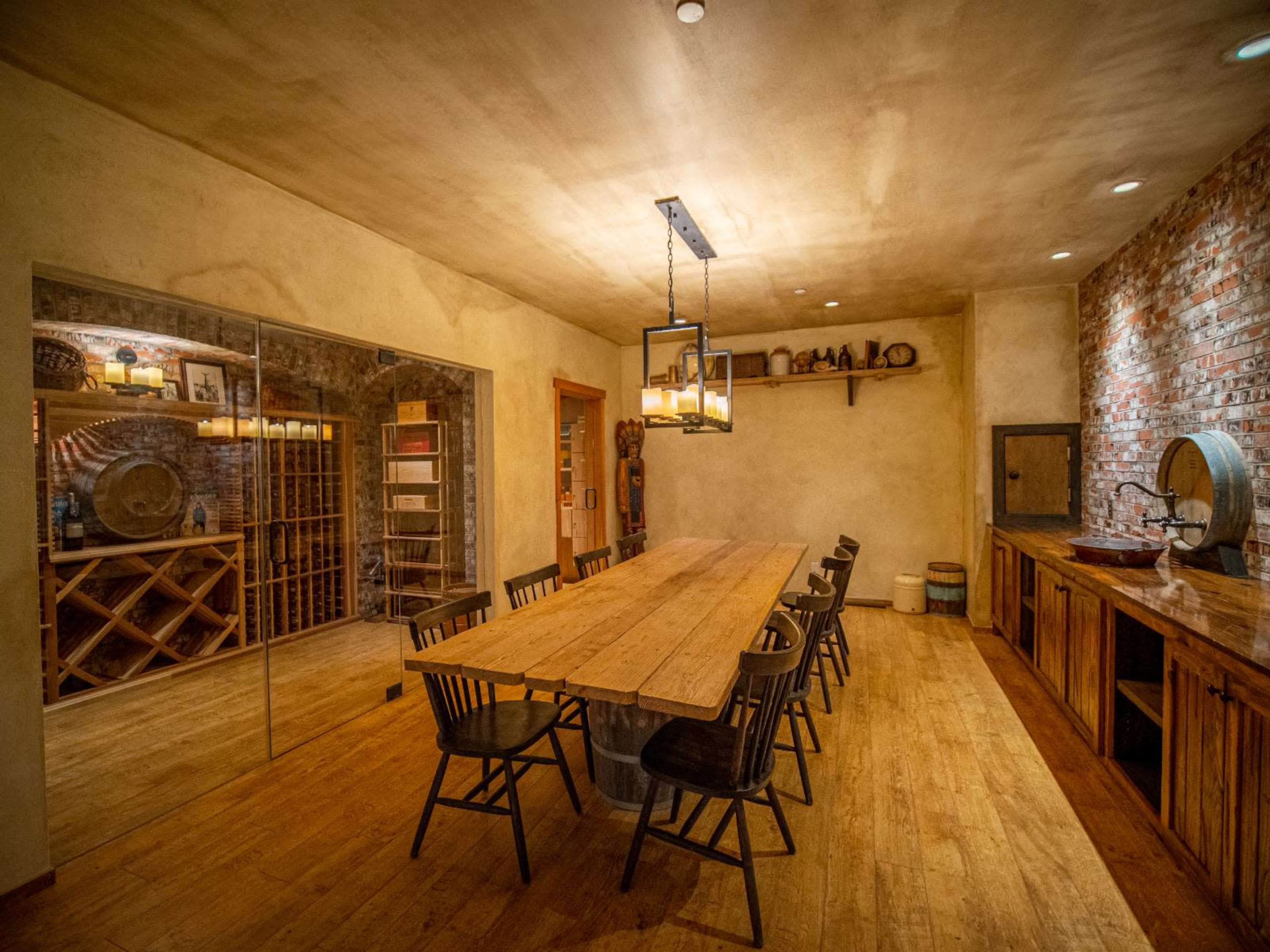 The image shows a dining room with a long wooden table surrounded by chairs, a wine rack on one side, and a rustic sink area on the other.