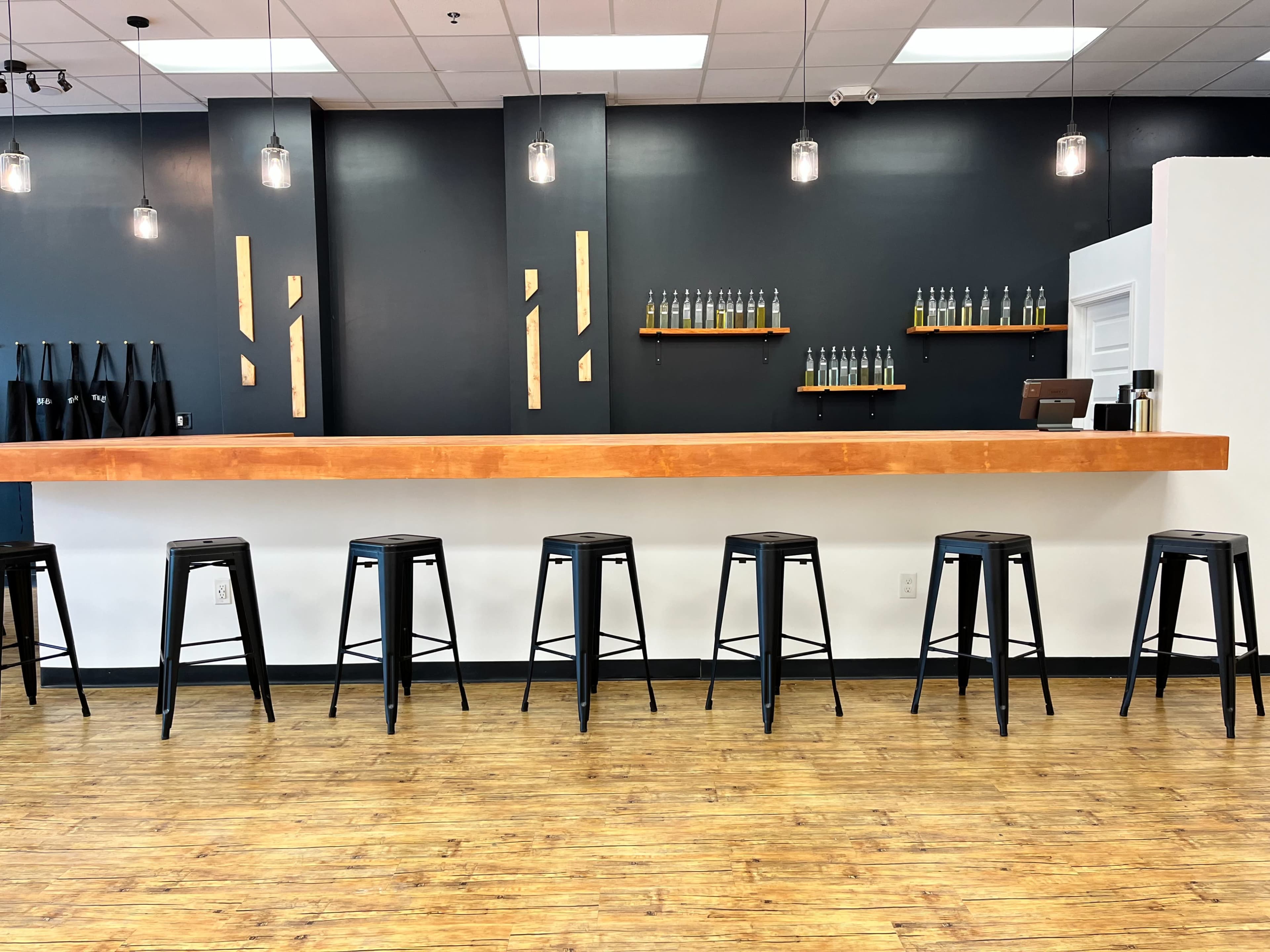 A minimalist bar area with a wooden countertop and several black stools lined up in front of a dark wall, featuring shelves with bottles on display.