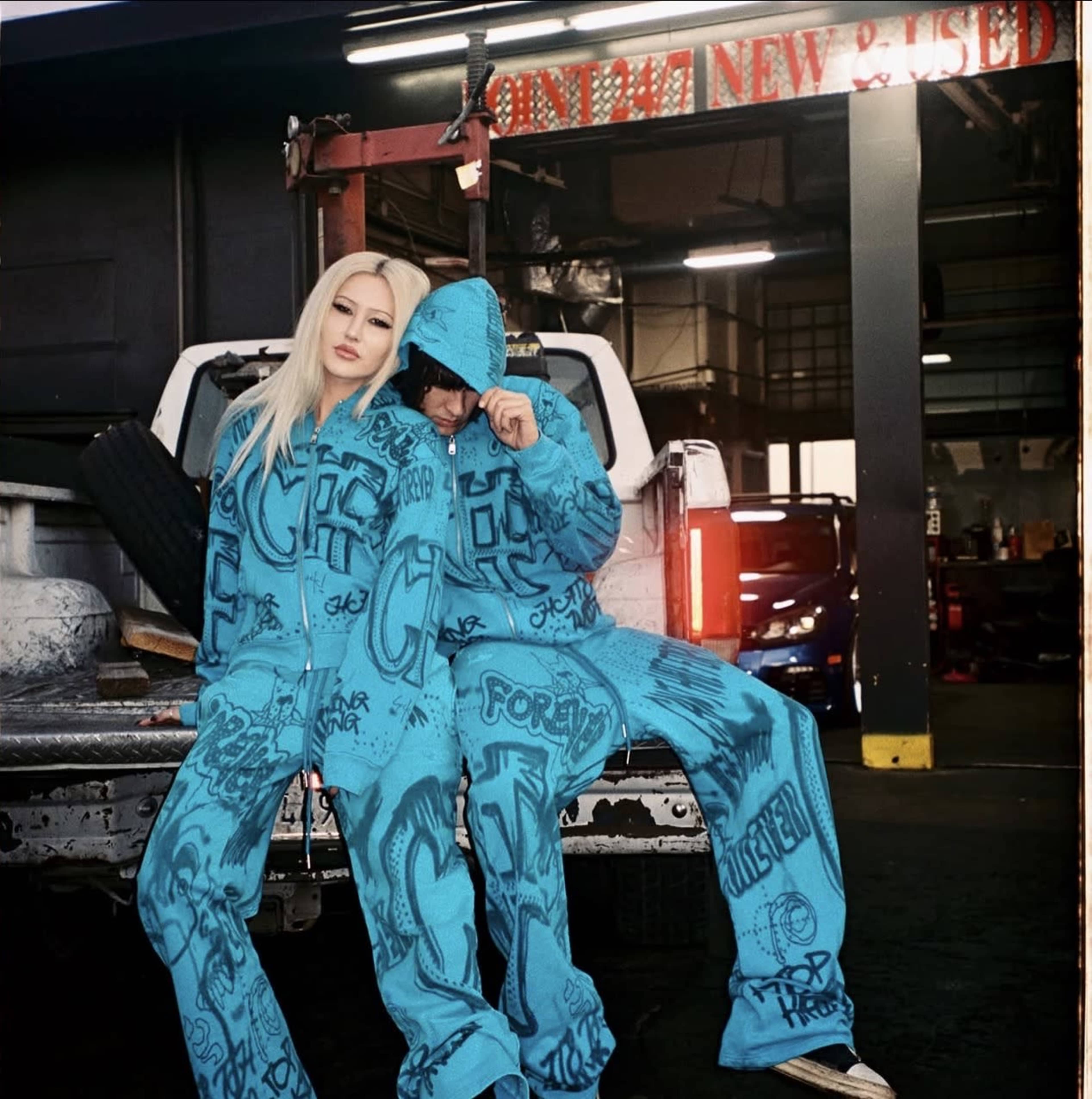 A man and woman wearing matching blue graffiti-patterned outfits pose on the back of a pickup truck in an industrial setting.