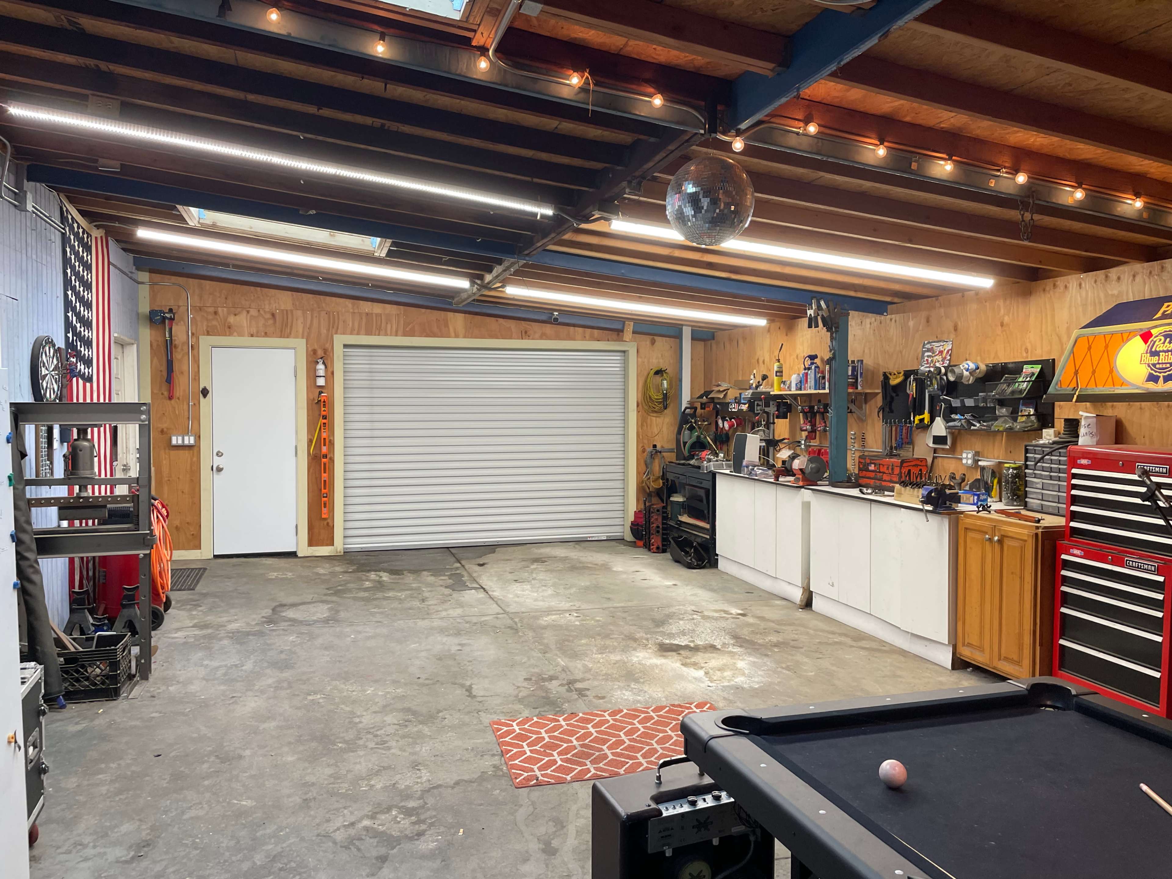 A spacious garage with wooden walls, a white door, a roll-up garage door, organized tool storage, and a pool table in the foreground.