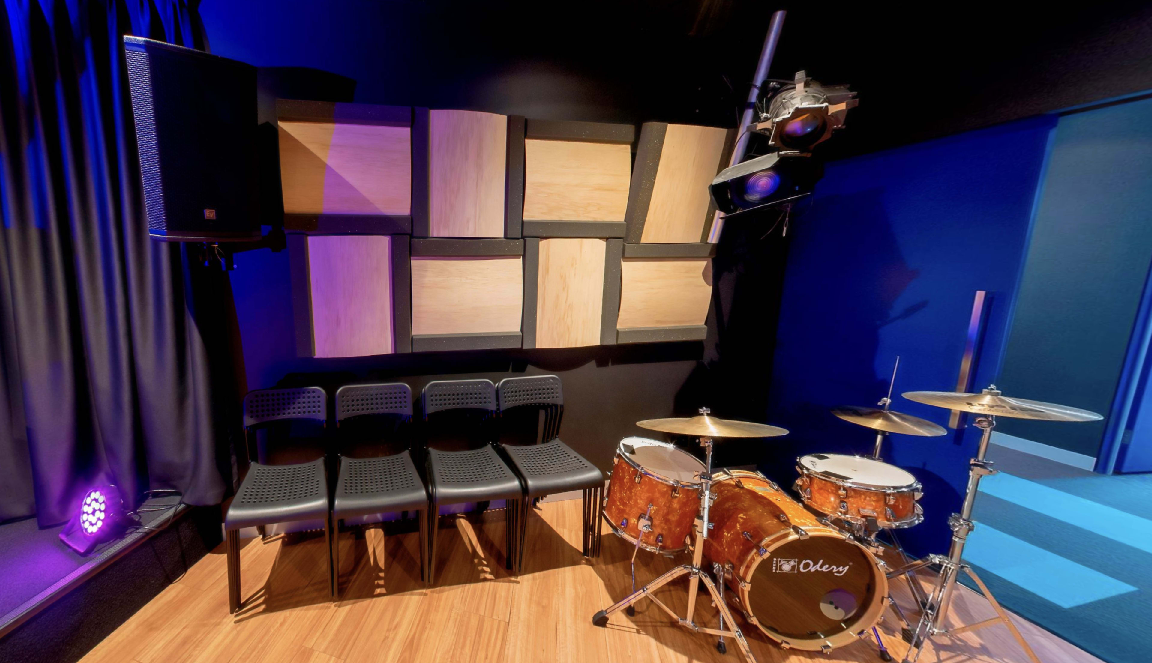 The image shows a music practice room with a drum set, a set of black chairs, and wooden sound panels on the wall.