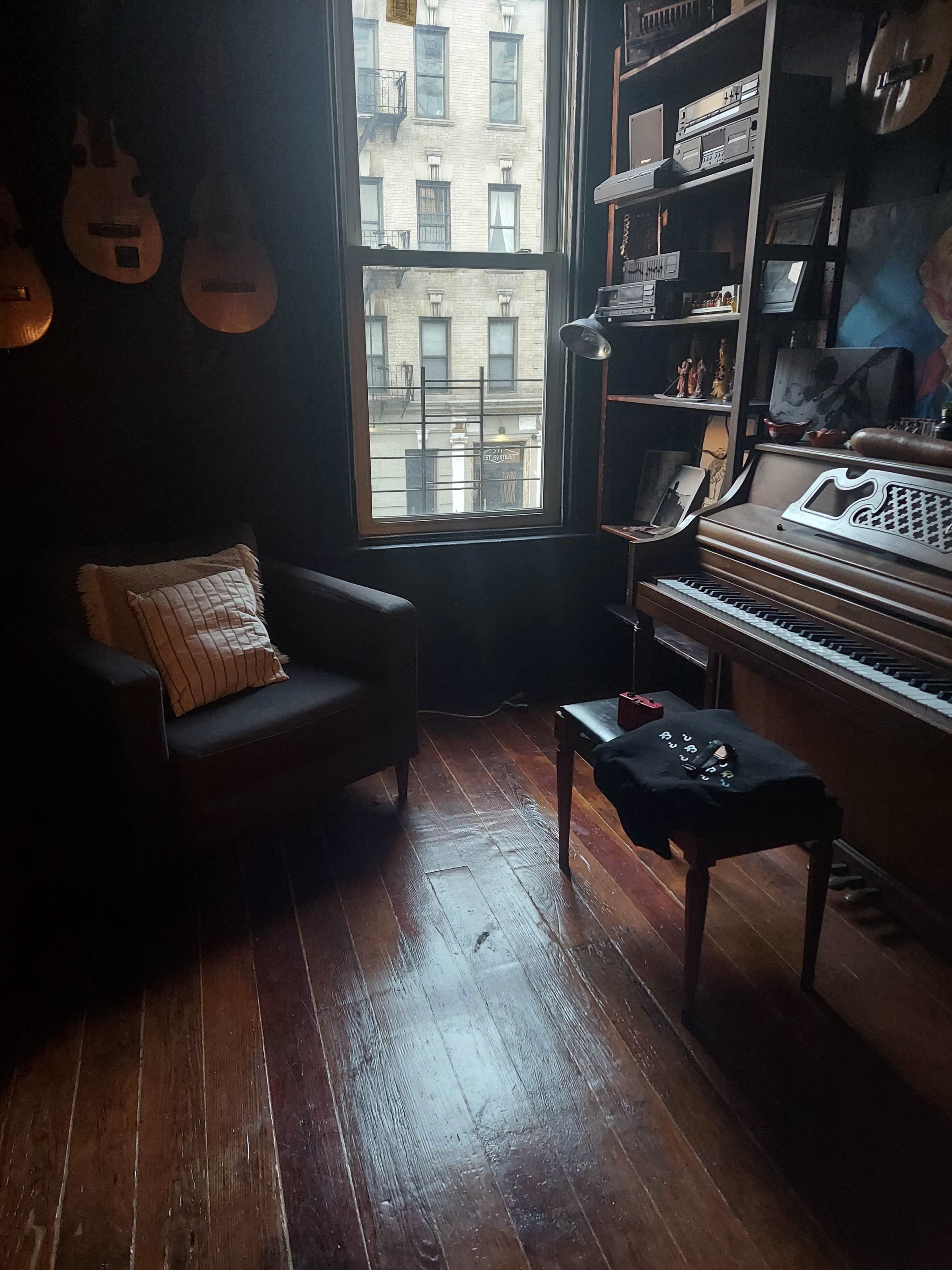 A cozy corner of a room features a chair next to a wooden piano and a small table with items on it, illuminated by natural light from a nearby window.