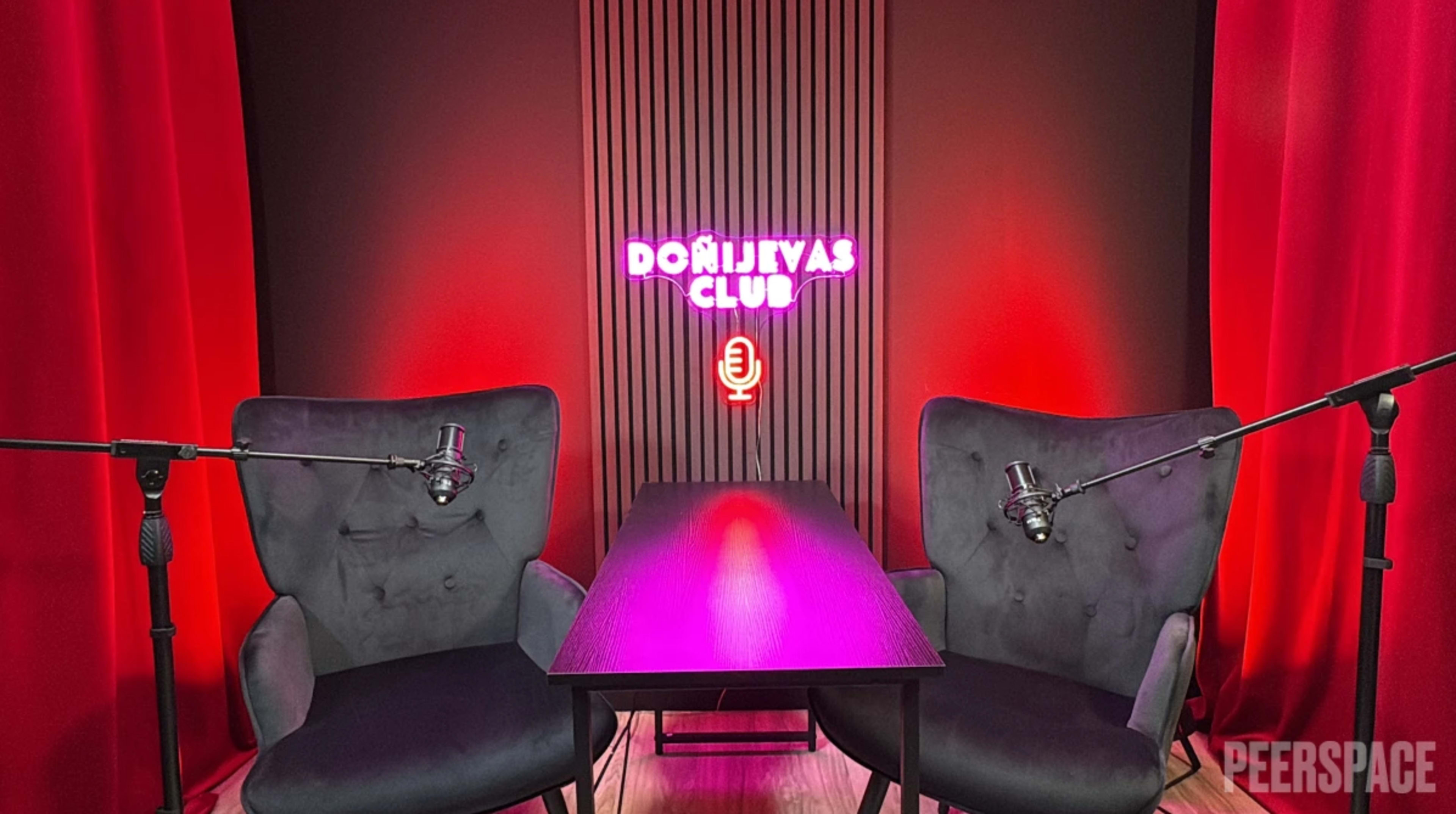 The image shows a podcast recording setup with two microphones positioned near two armchairs facing a small table, illuminated by a neon sign that reads "DONIJIVAS CLUB."