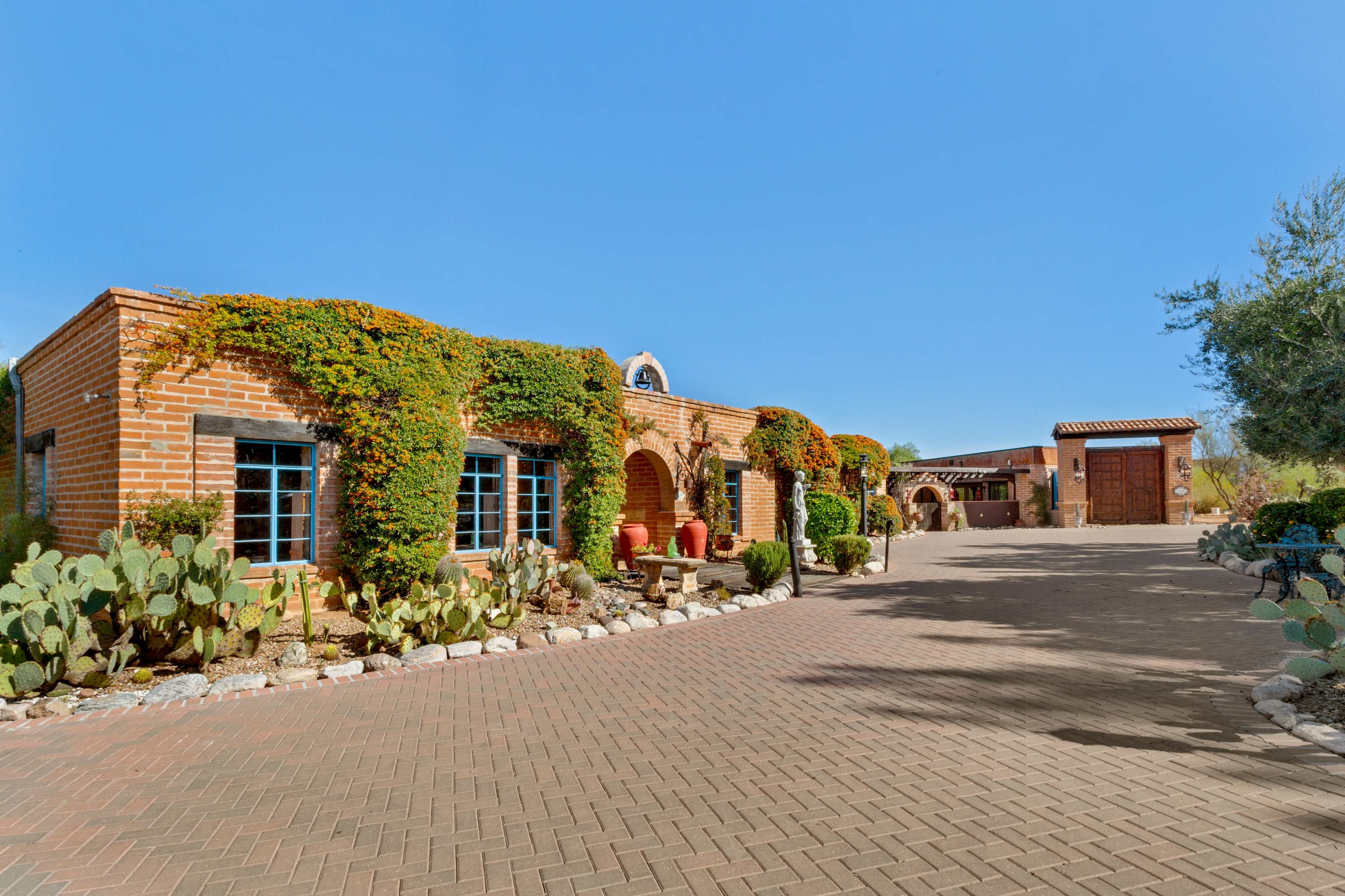 A brick house adorned with climbing plants is situated beside a paved driveway that leads to a separate wooden garage in a clear blue sky.