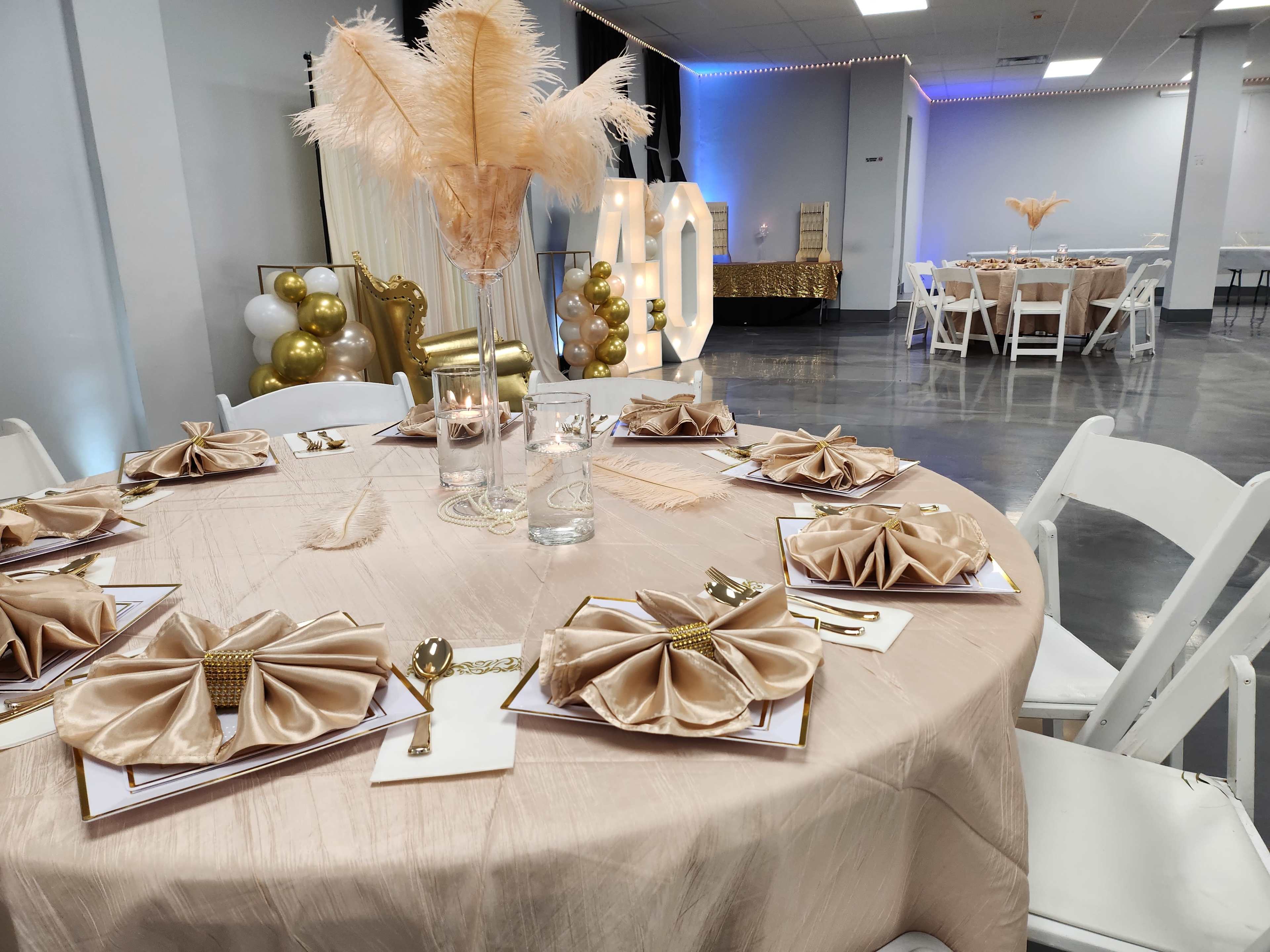 A banquet hall is set up with elegantly arranged tables featuring gold and beige decor, feather centerpieces, and neatly folded napkins.