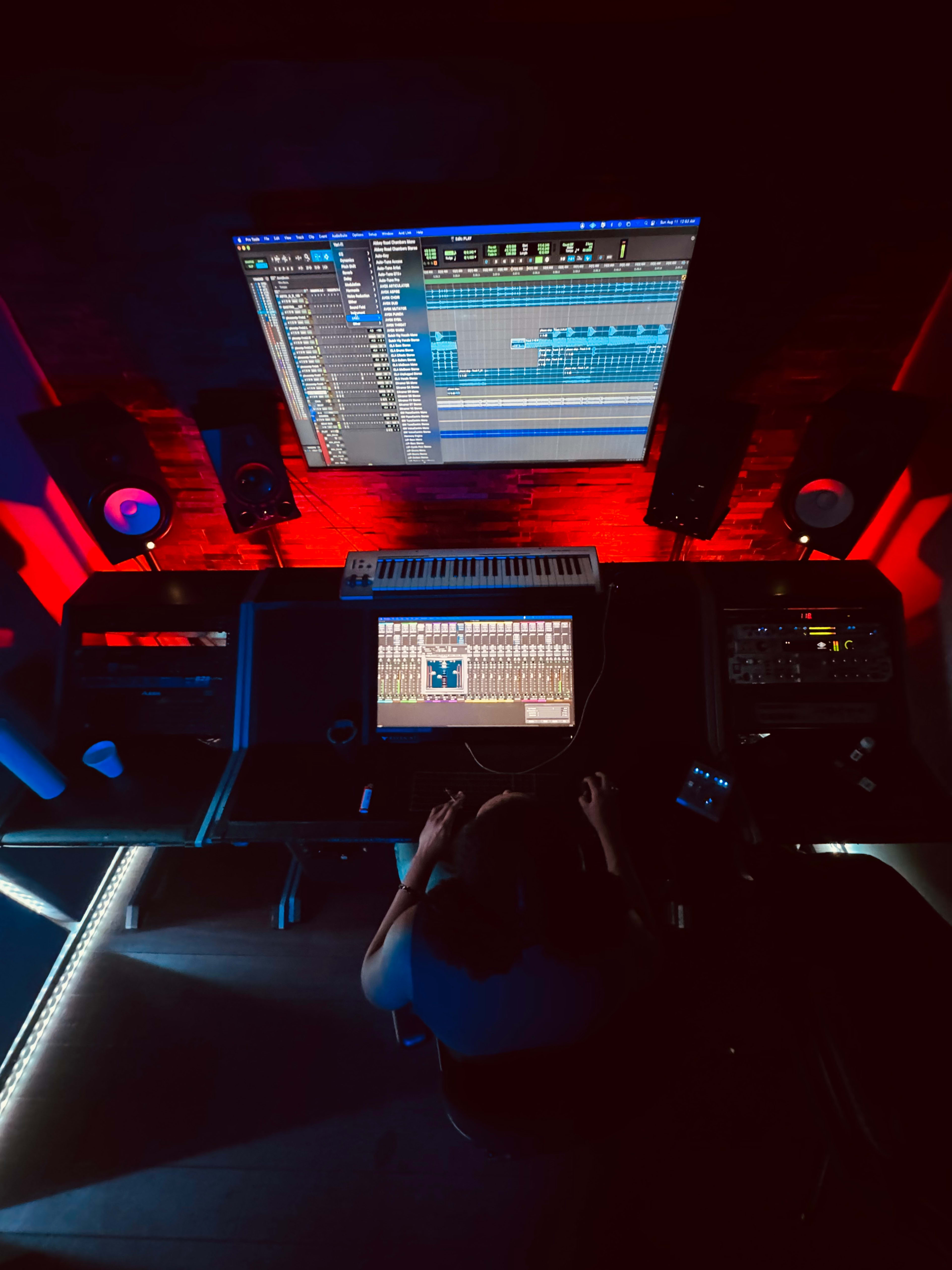 A person works at a music production studio with a computer, keyboard, and sound equipment illuminated by red lighting.