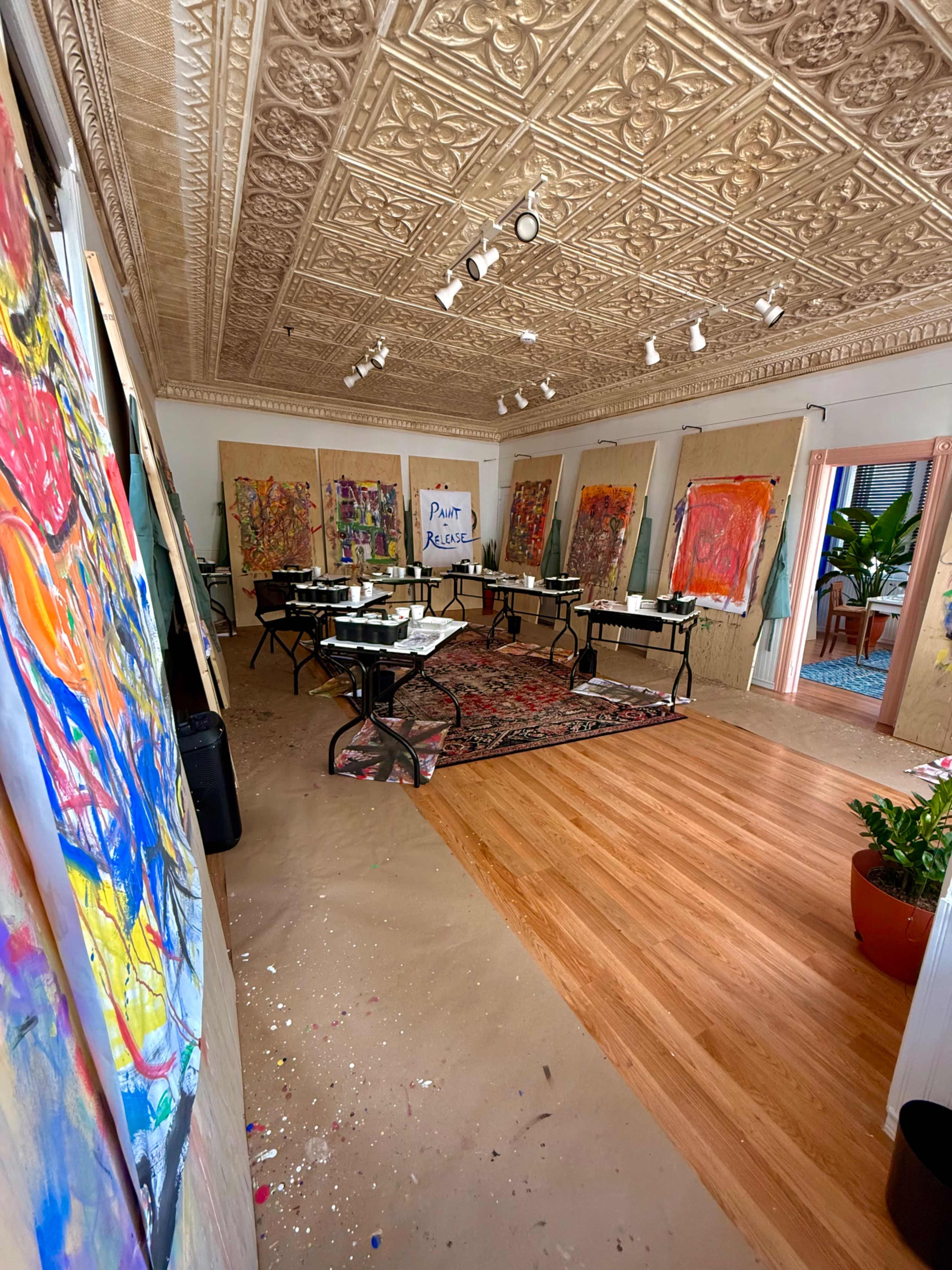 The image shows an art studio featuring colorful paintings on display, a large table in the center, and a decorative ceiling.