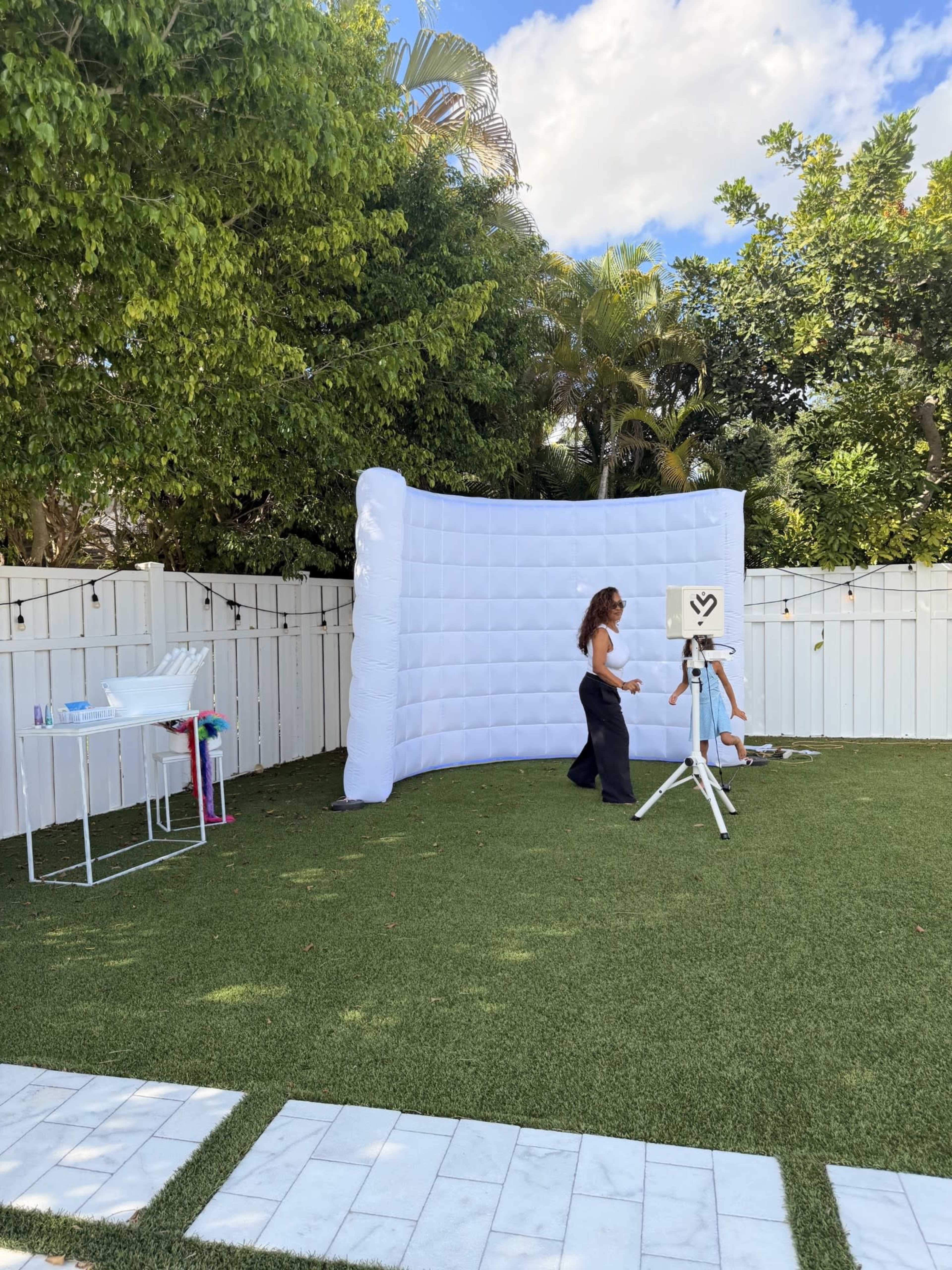 A woman stands near an inflatable white backdrop in a backyard, which is surrounded by greenery and features a small table nearby.