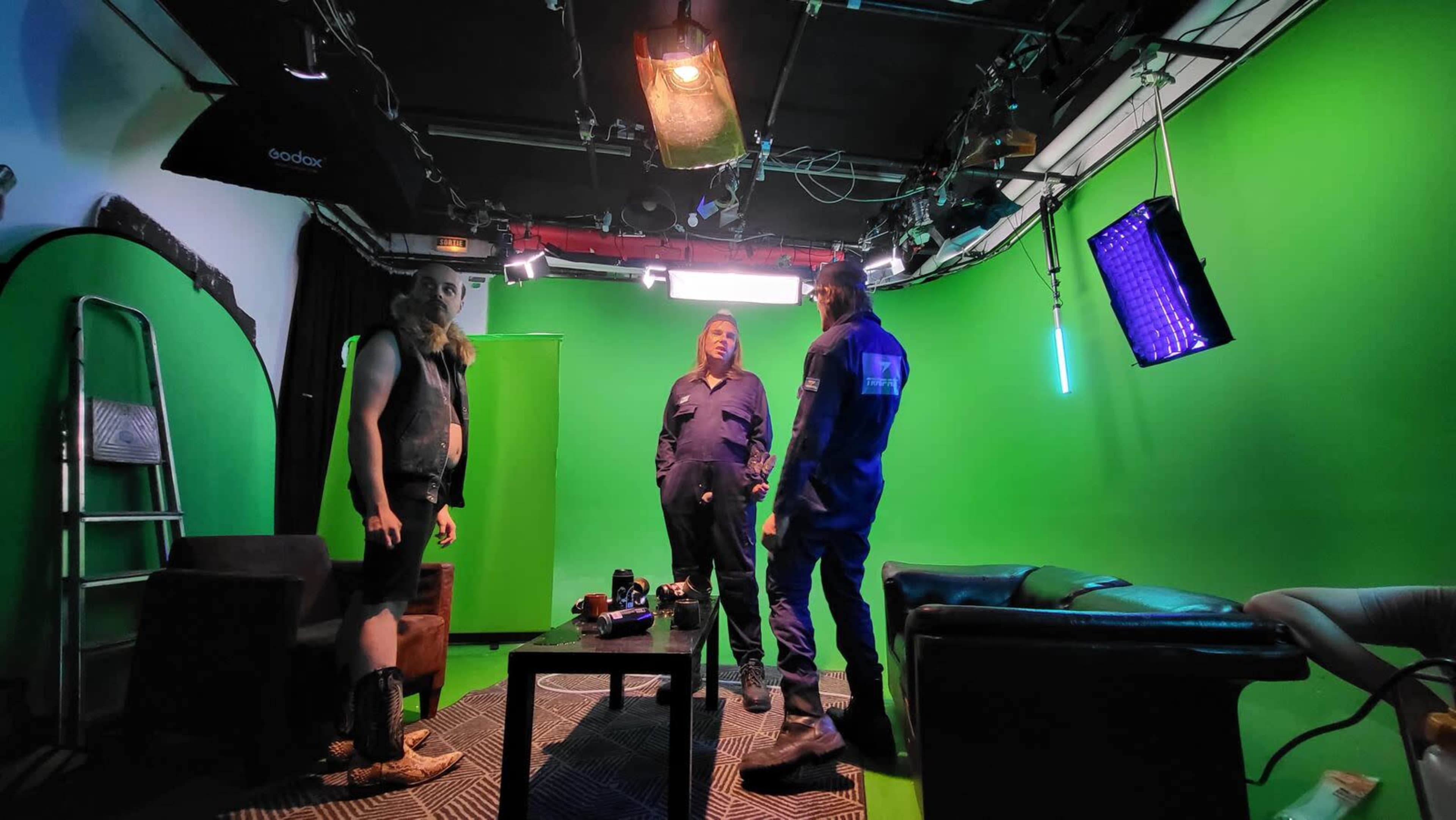 Three individuals are positioned in a studio with a green screen, surrounded by studio equipment and furniture.