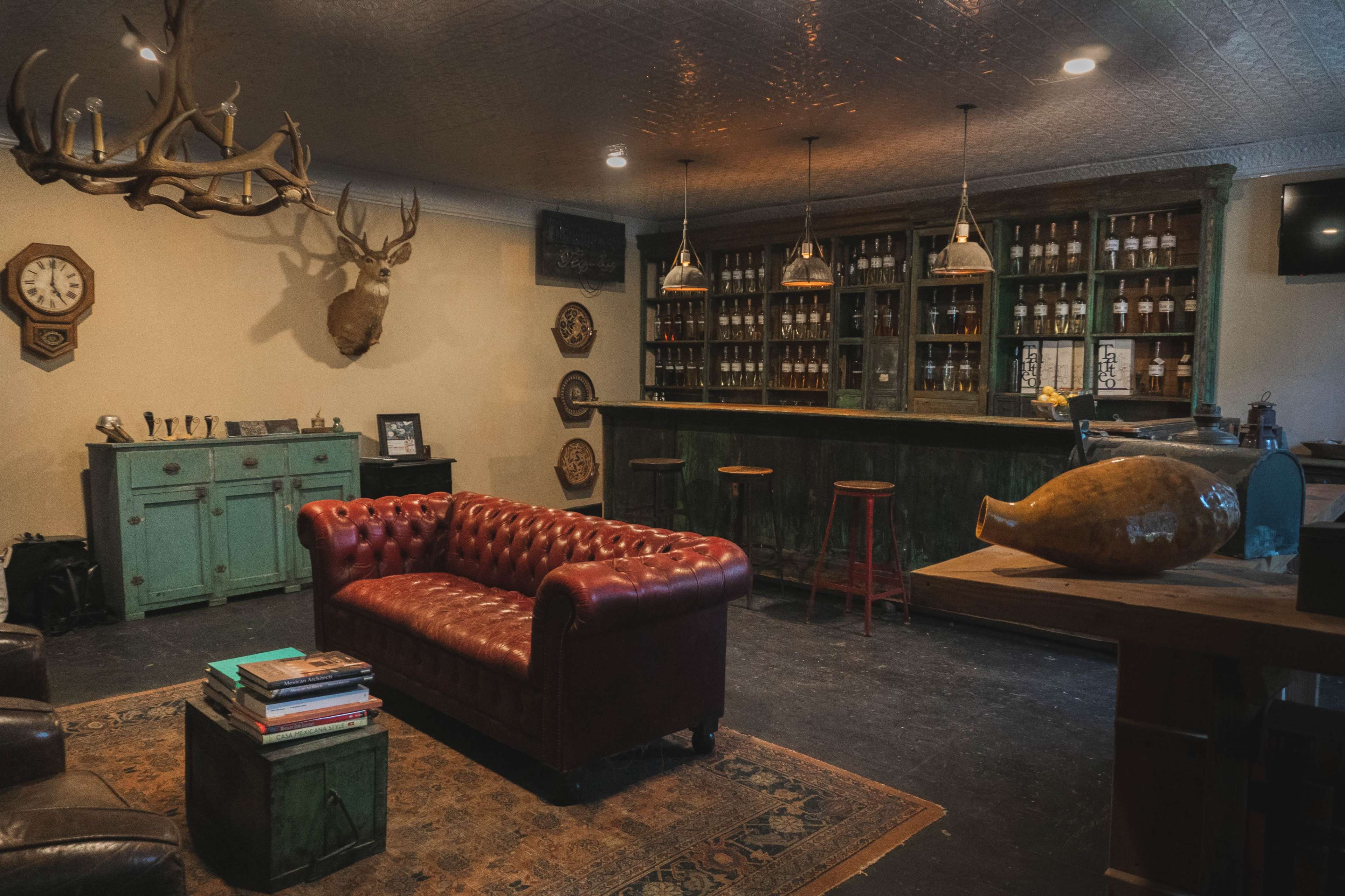 A cozy bar-style room featuring a red leather chesterfield sofa, a wooden bar stocked with bottles, and rustic decor including antlers and a clock on the wall.