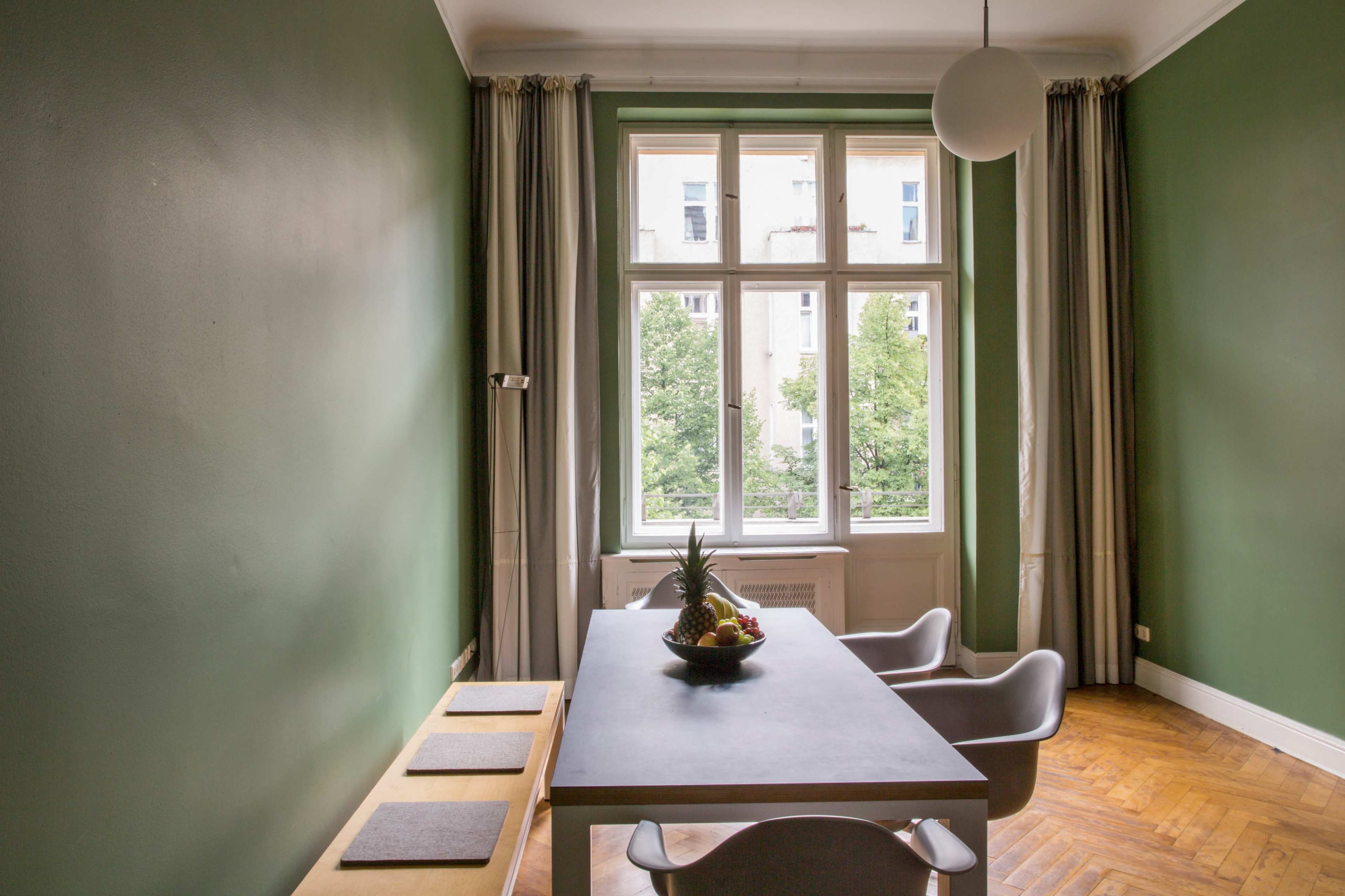 A dining area features a table with a fruit bowl, surrounded by modern chairs and large windows overlooking a green outdoor area.
