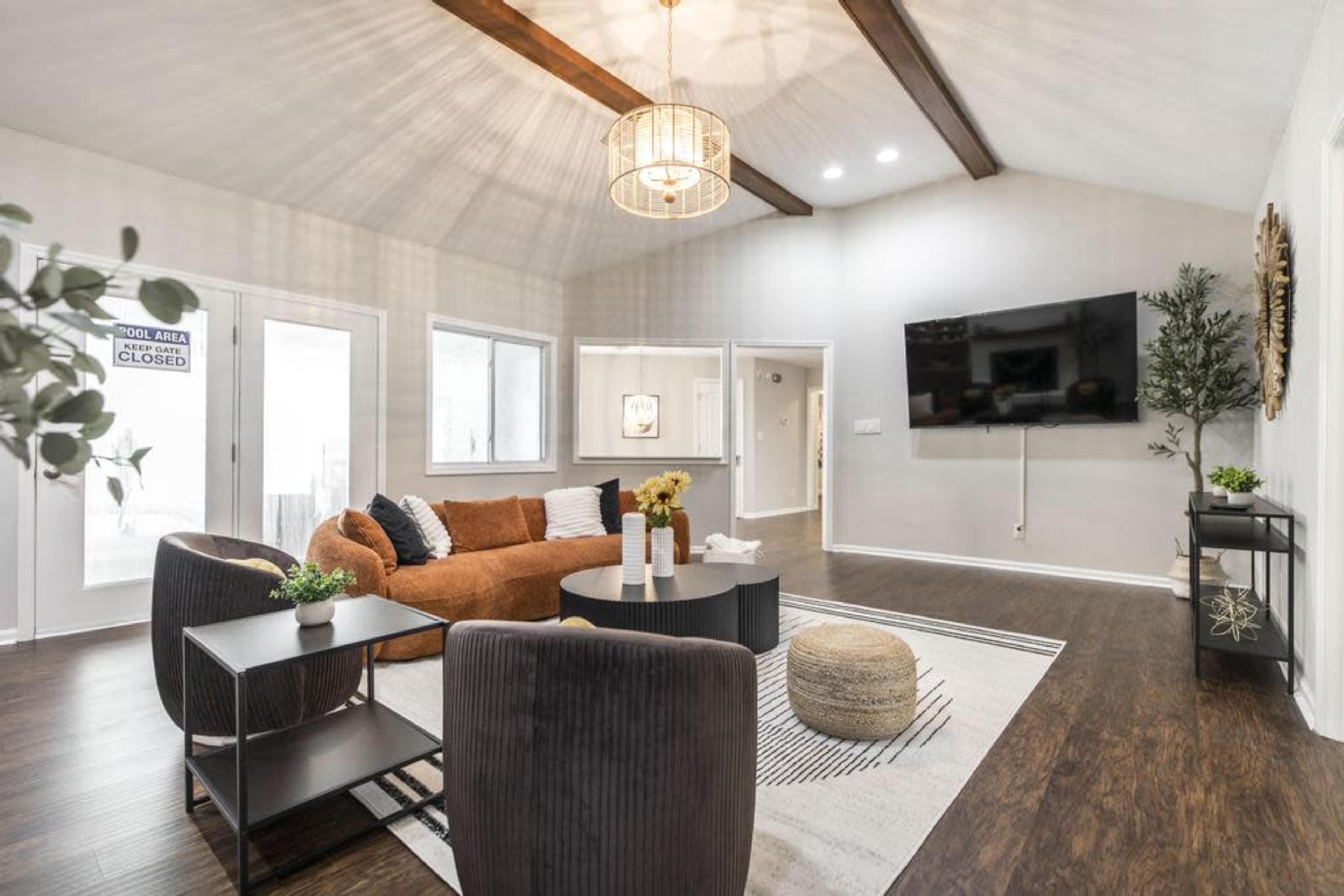 A modern living room features a brown sofa, a black coffee table, and two black chairs, with a large TV mounted on the wall and natural light coming through the windows.