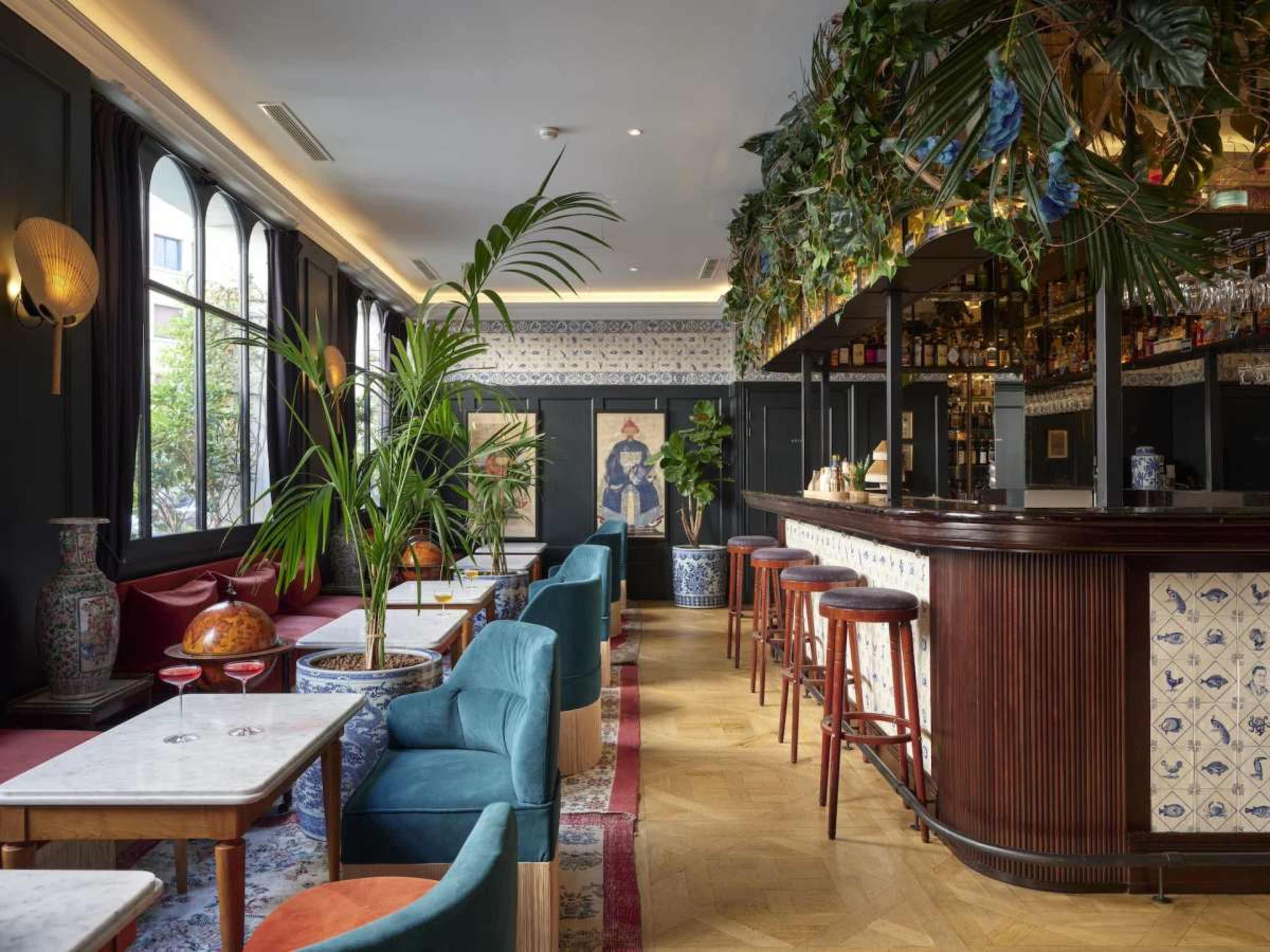 The image shows a stylish bar interior featuring colorful armchairs, a wooden bar counter, and tropical plants, with a decorative mural and tiled accents on the walls.
