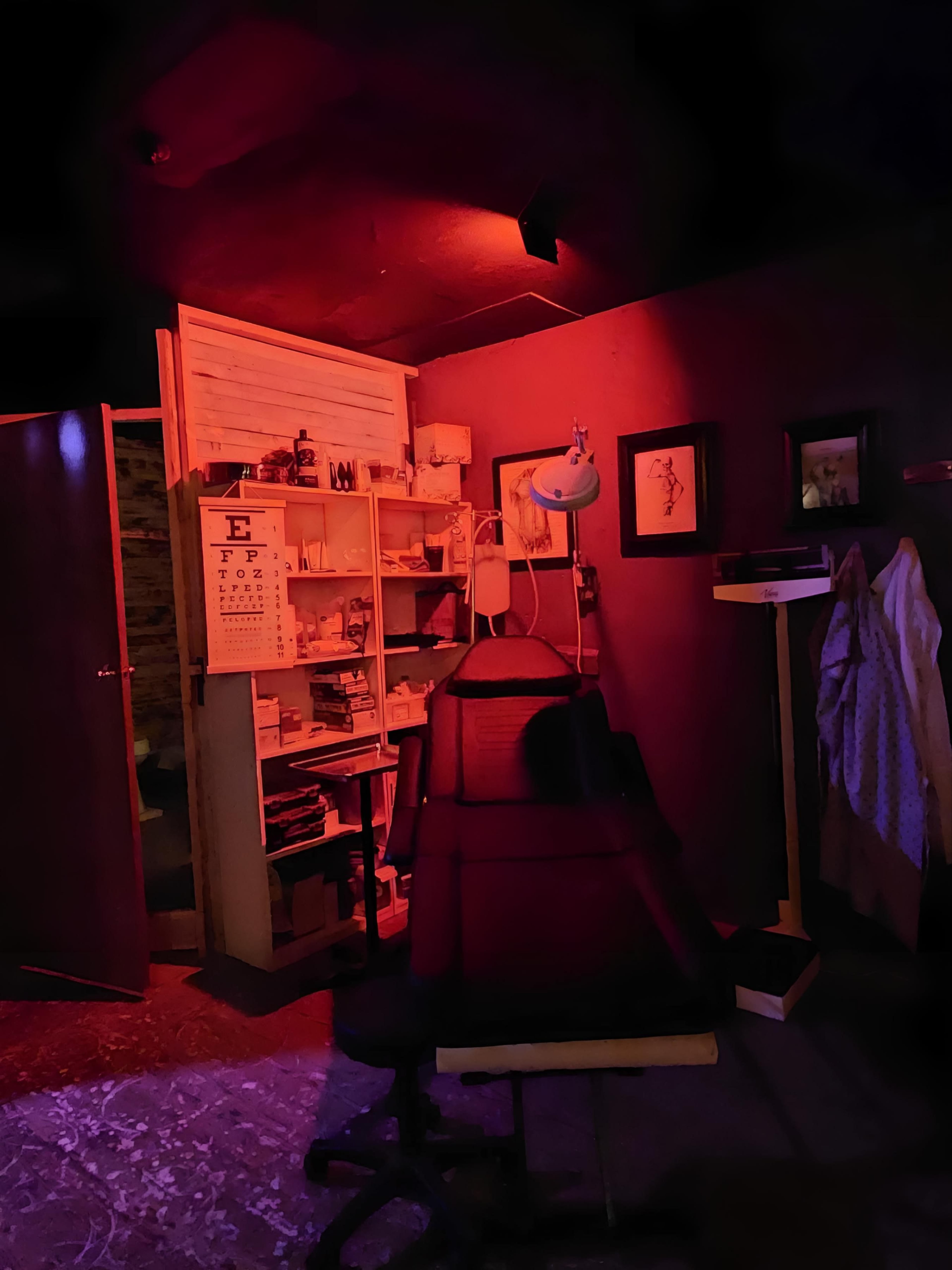 A dimly lit room features a dental chair, a bookshelf filled with items, and an eye chart on the wall.