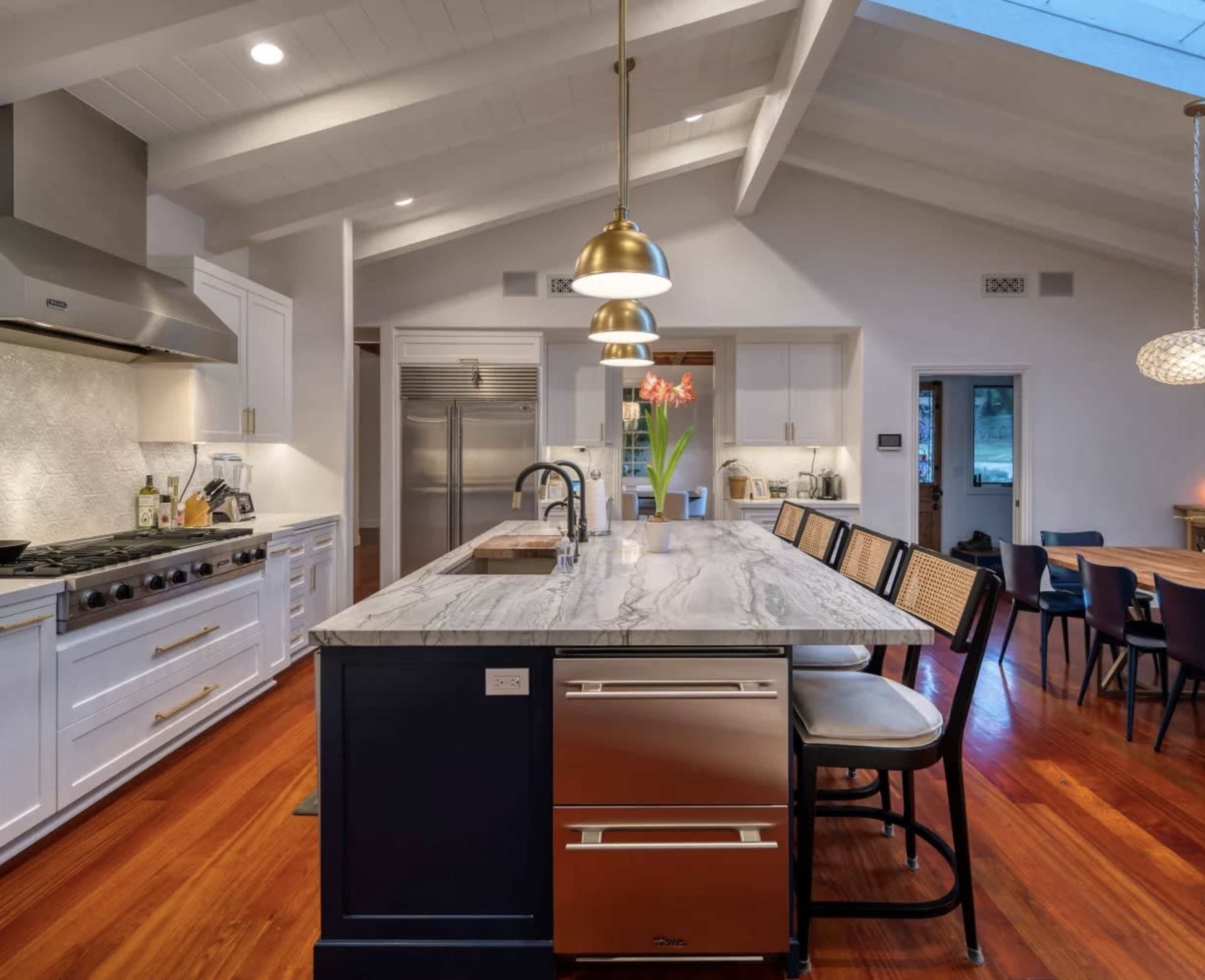 The image shows a modern kitchen featuring a large island with a marble countertop, stainless steel appliances, and wooden beams on the ceiling.