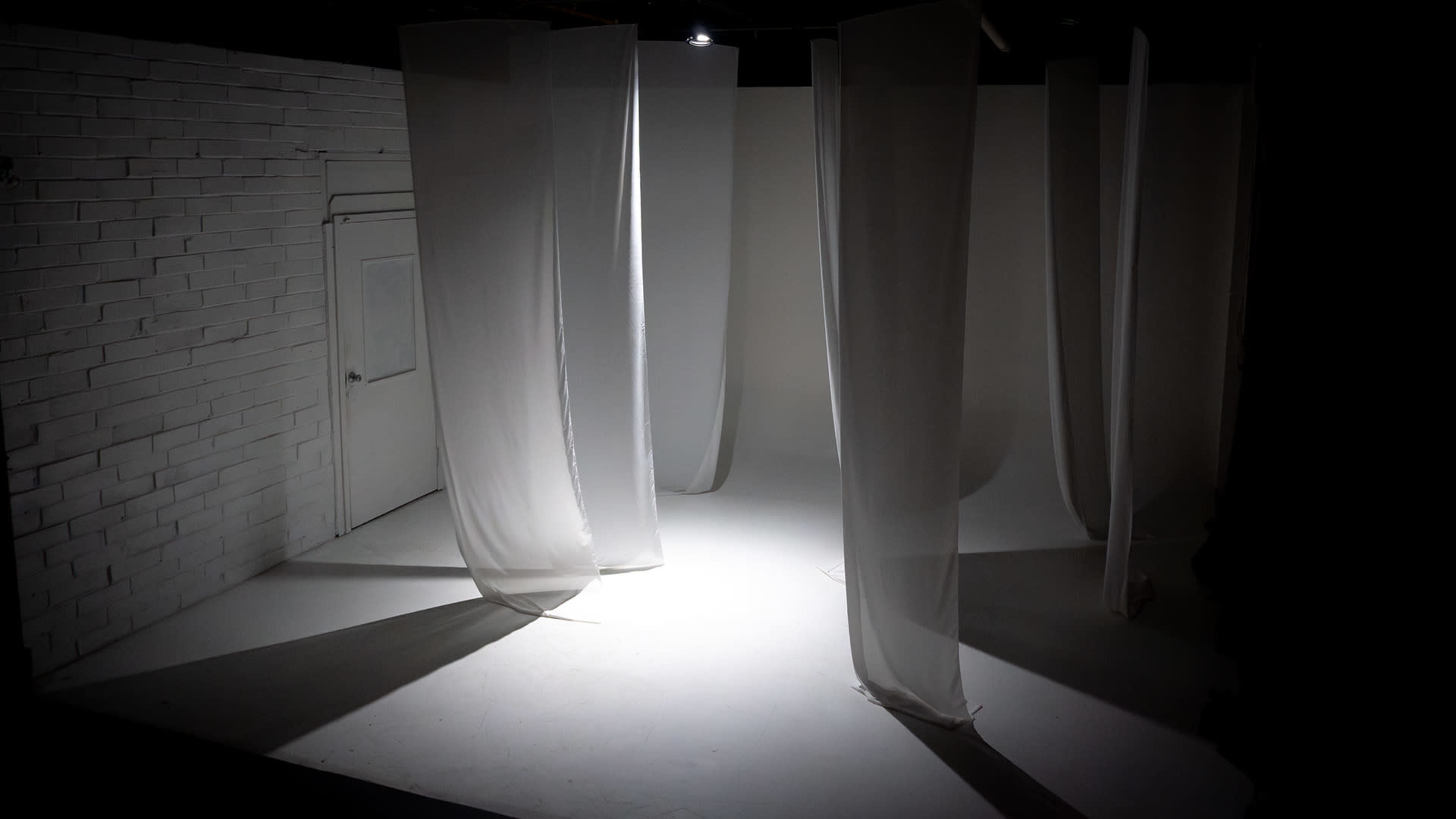 The image shows a minimalist interior space with white walls and several sheer fabric panels hanging from the ceiling, casting shadows on the floor.